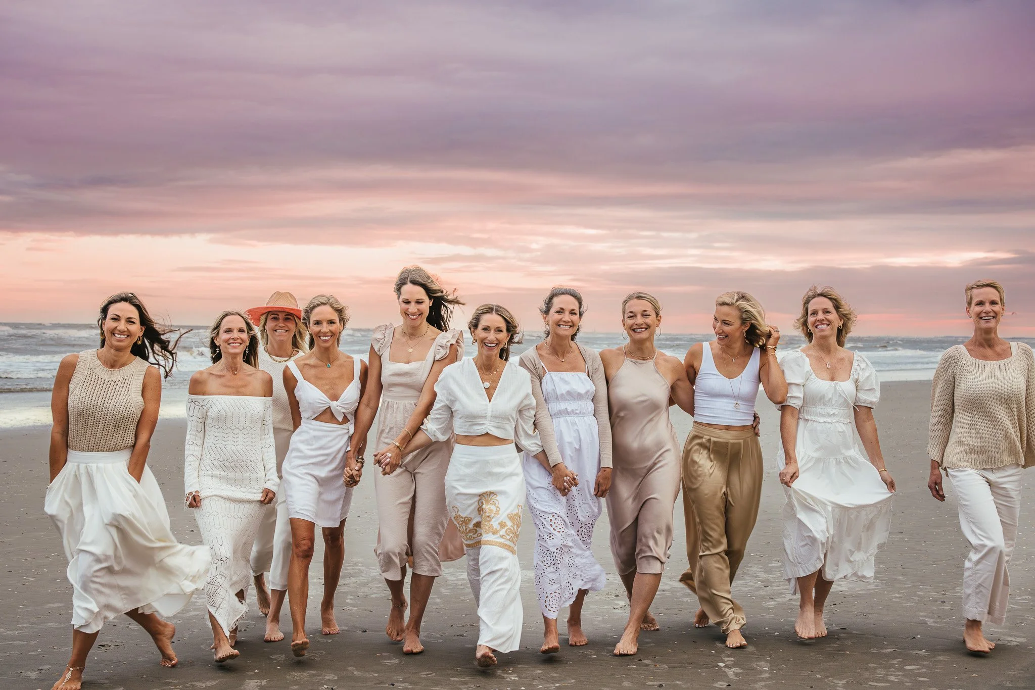 A group of women in white and beige clothing walking on the beach during sunset, holding hands and smiling.