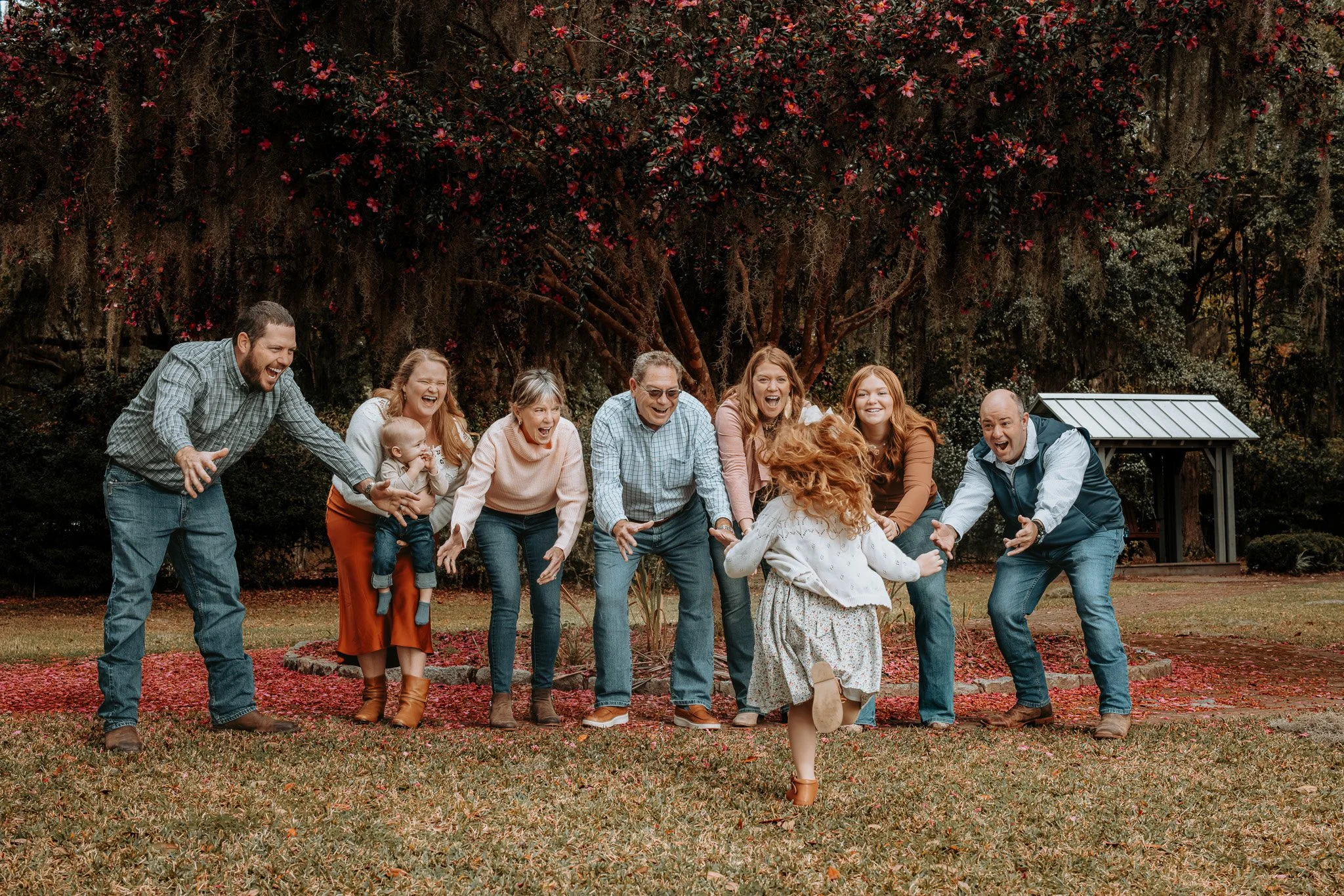 Group of eight people, including children, playing and running in a park with trees and blooming flowers in the background.
