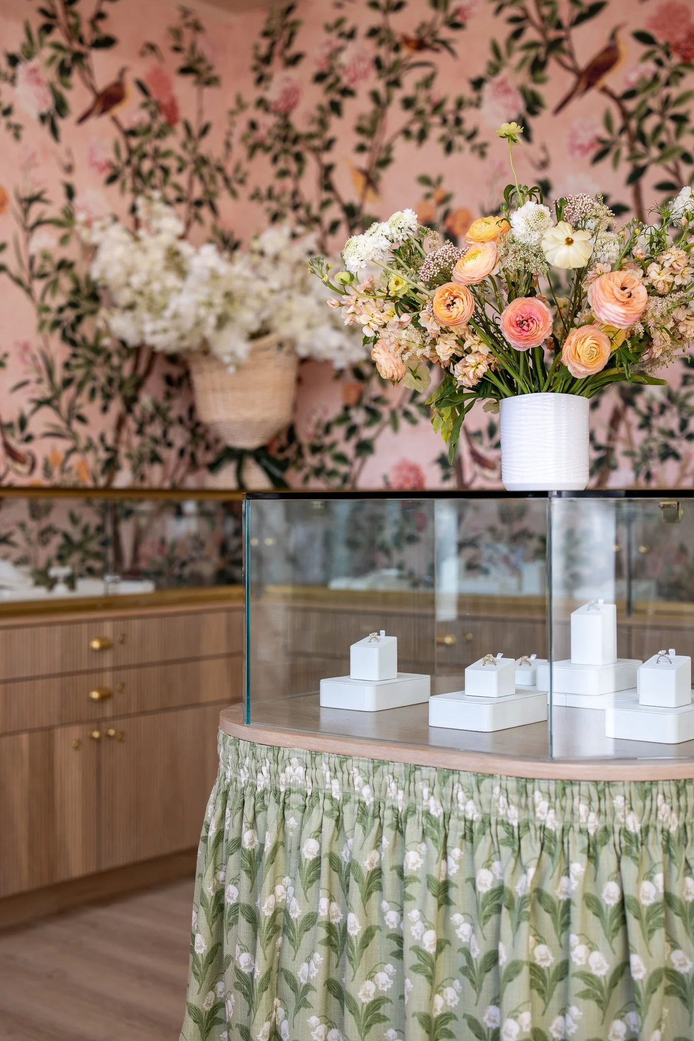 A display of rings in a jewelry store with a bouquet of pink and cream flowers on top of a glass case, floral wallpaper, and wooden cabinetry in the background.