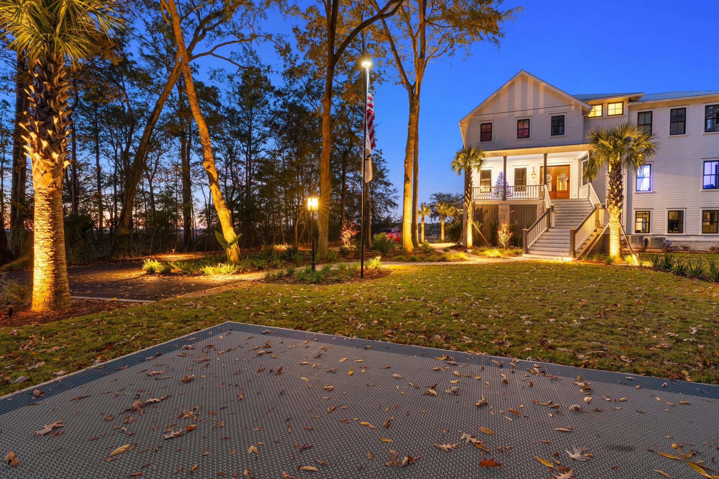 A large white house with a front porch and stairs, illuminated at dusk, with palm trees and outdoor lighting. The sky is a deep blue, and the ground has some fallen leaves. There are street lamps and an American flag in front of the house.
