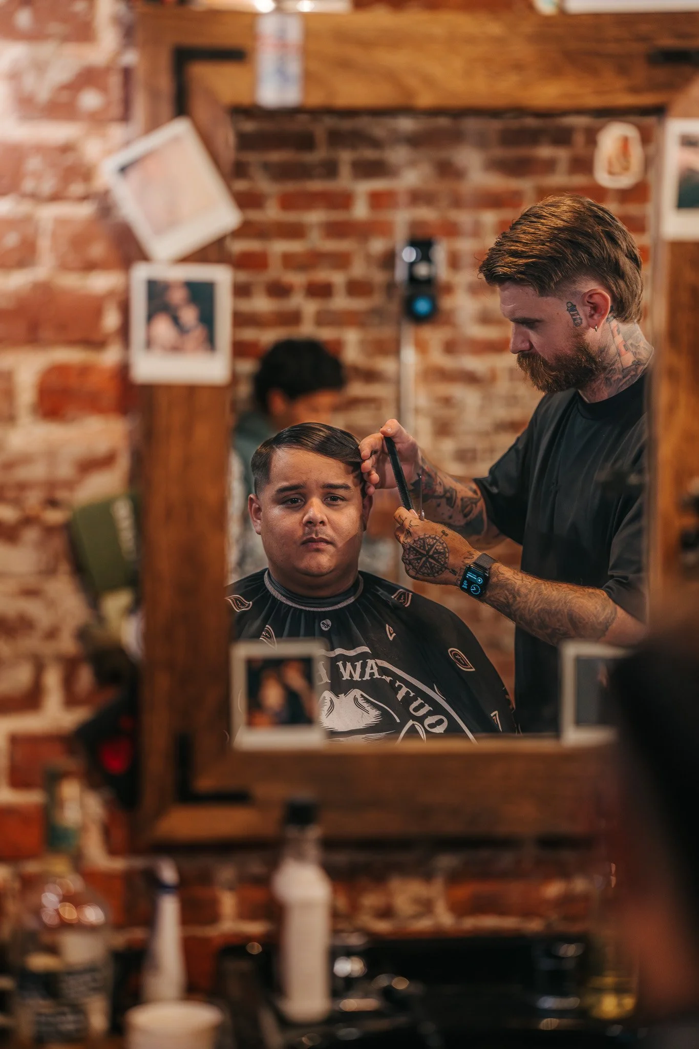 A man with tattoos getting a haircut from a barber with tattoos in a barbershop with a brick wall, seen through a mirror.