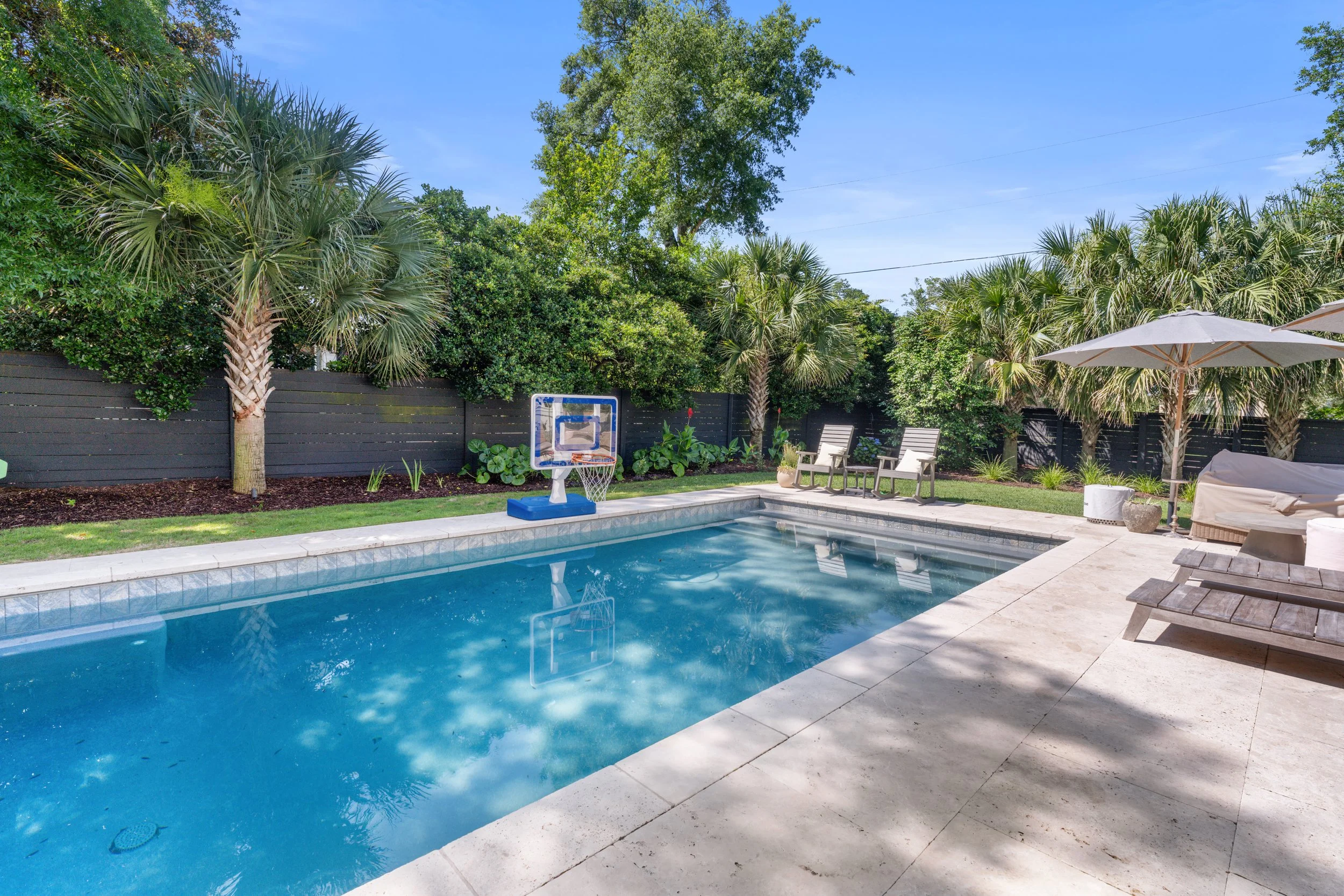 Residential backyard with swimming pool, palm trees, outdoor furniture, umbrellas, and a black fence, under a clear blue sky.