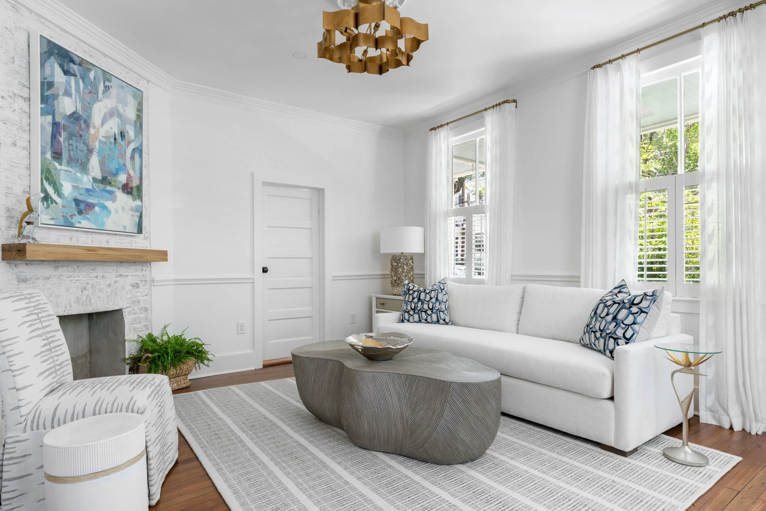 Bright living room with white walls, large windows with white curtains, white sofa with blue patterned pillows, round textured coffee table, armchair with striped fabric, potted fern, abstract painting above fireplace, lamp with textured base, decora
