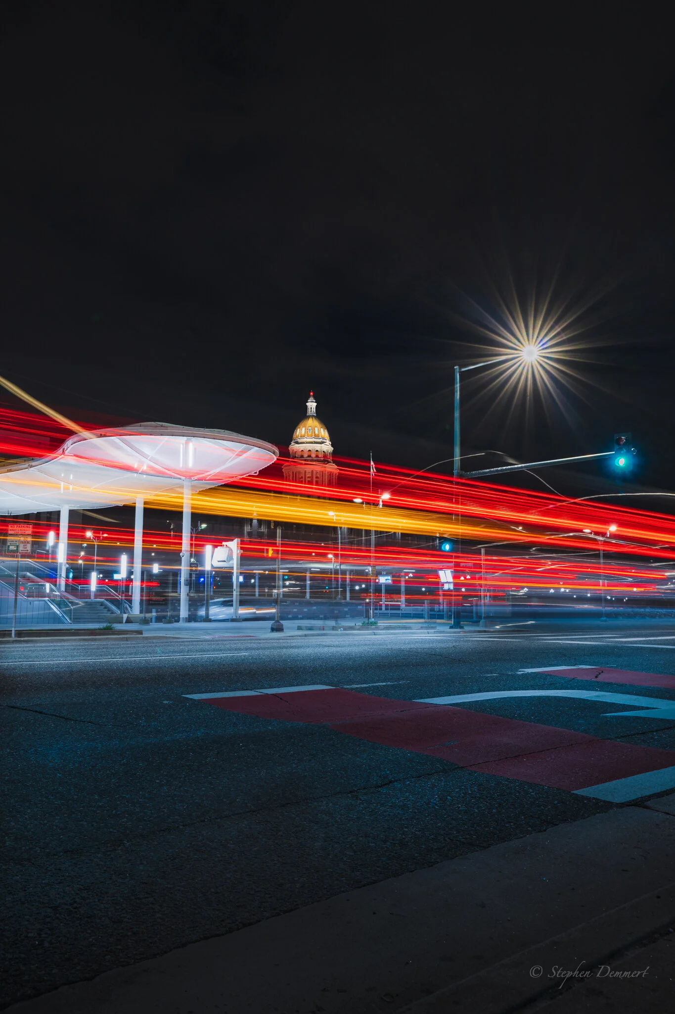 Nighttime city street with light trails from moving vehicles, a historic domed government building in the background, and a streetlamp emitting bright light. Long exposure photography captures motion and illuminated street elements.