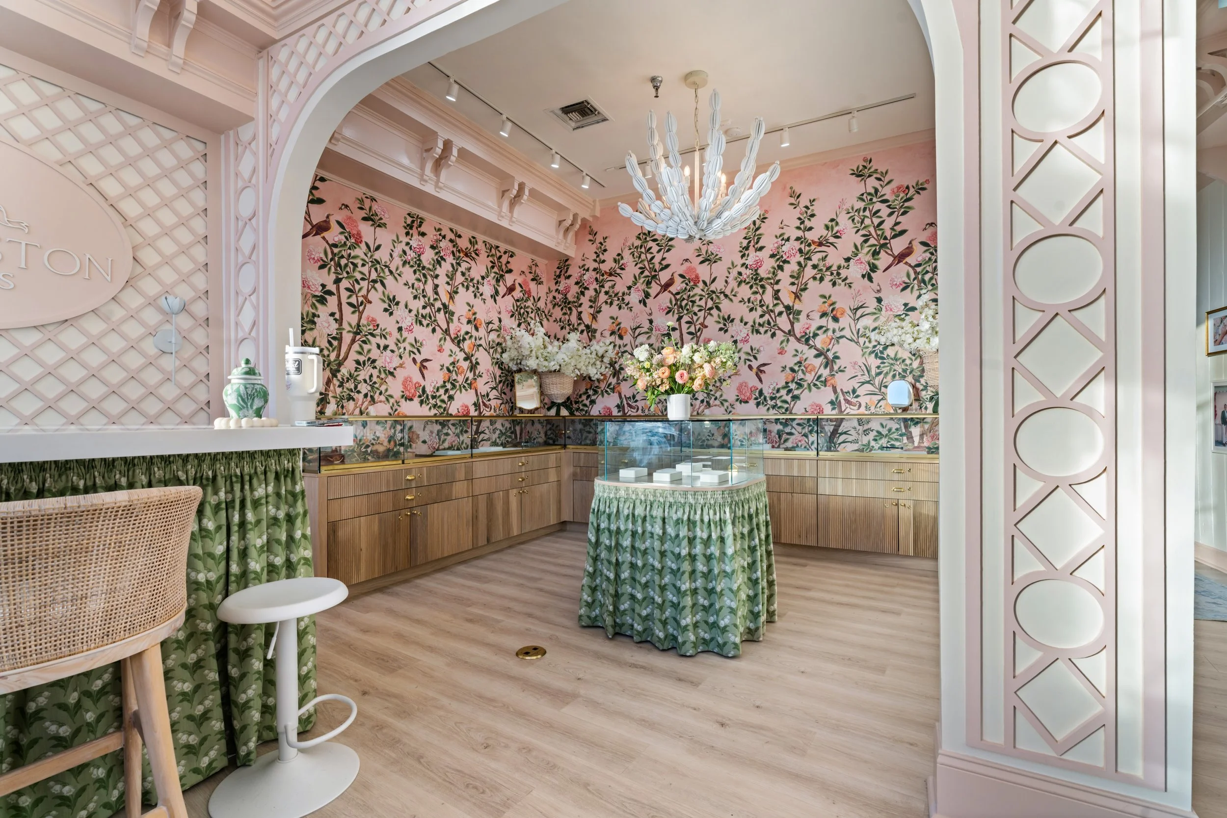 Interior of a boutique store with a pink floral wallpaper, wooden cabinetry, glass display case, floral arrangements, chandelier, and decorative pink and green accents.