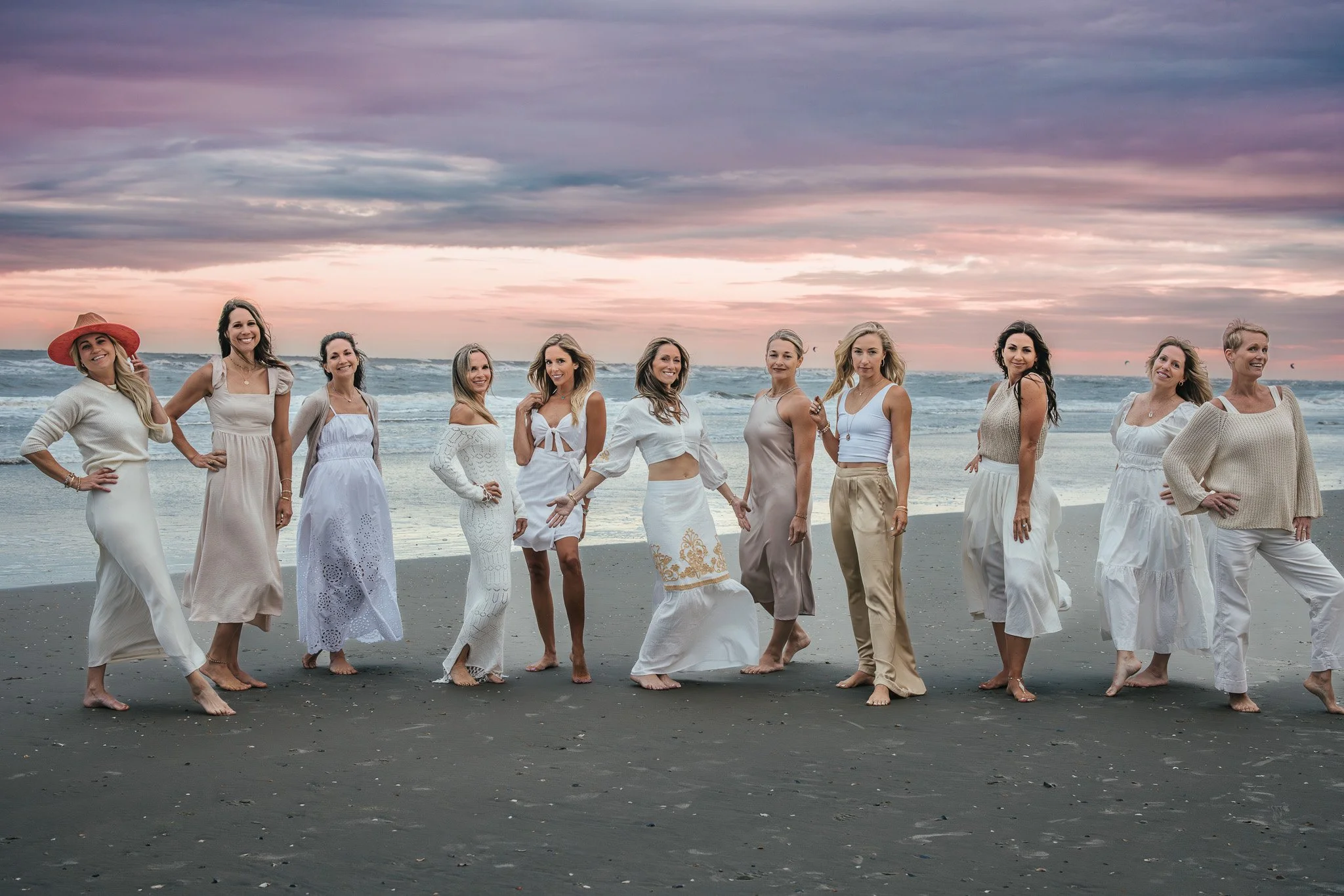 Group of women in white and beige dresses and casual clothing standing on a beach at sunset, smiling and posing.