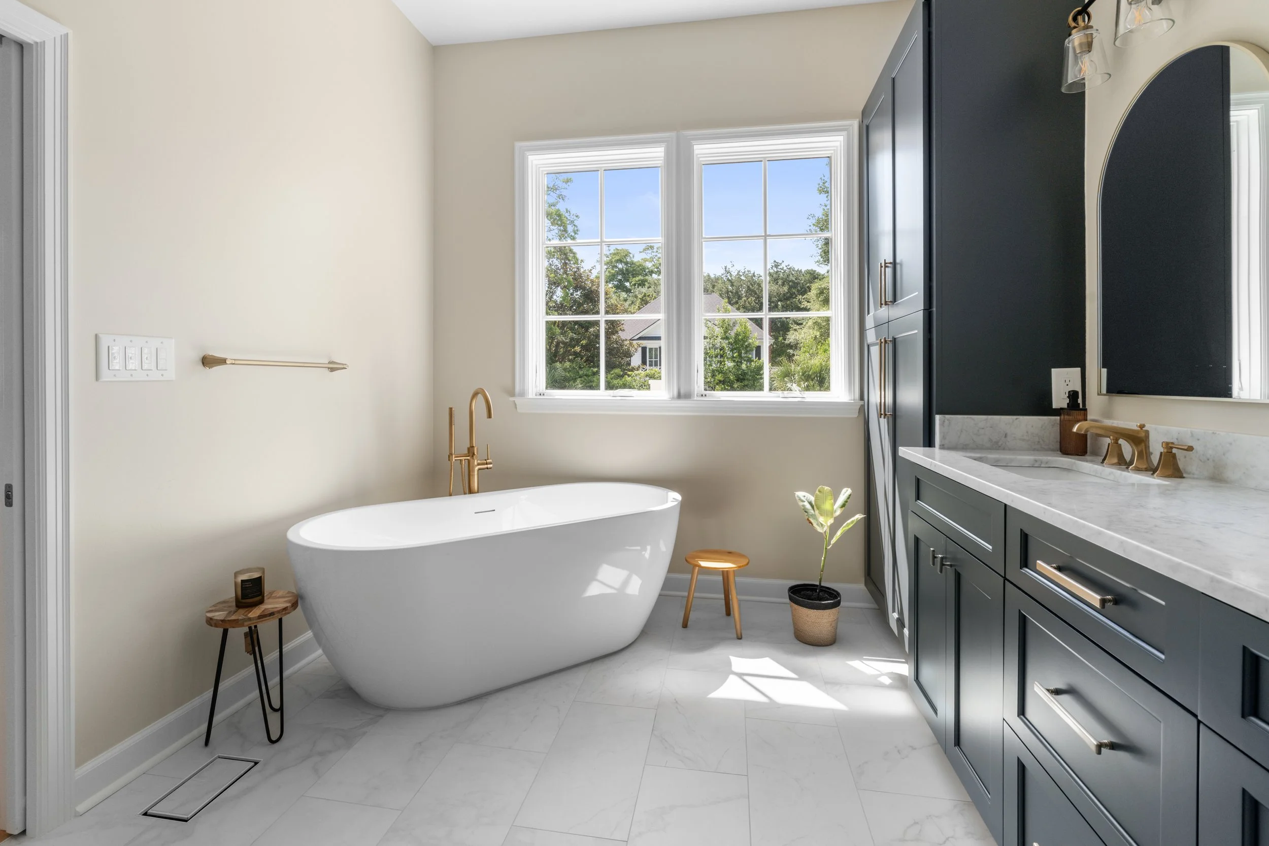 Modern bathroom with a white bathtub, gold fixtures, a navy blue vanity with a marble countertop, and a large window with a view of greenery.
