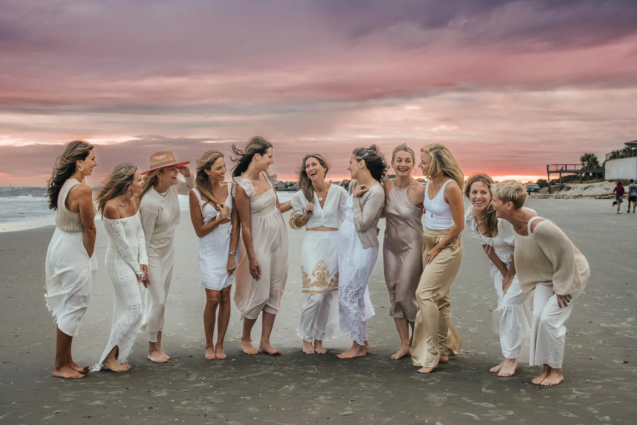 Group of women dressed in white and beige clothing, laughing and talking on a beach during sunset.