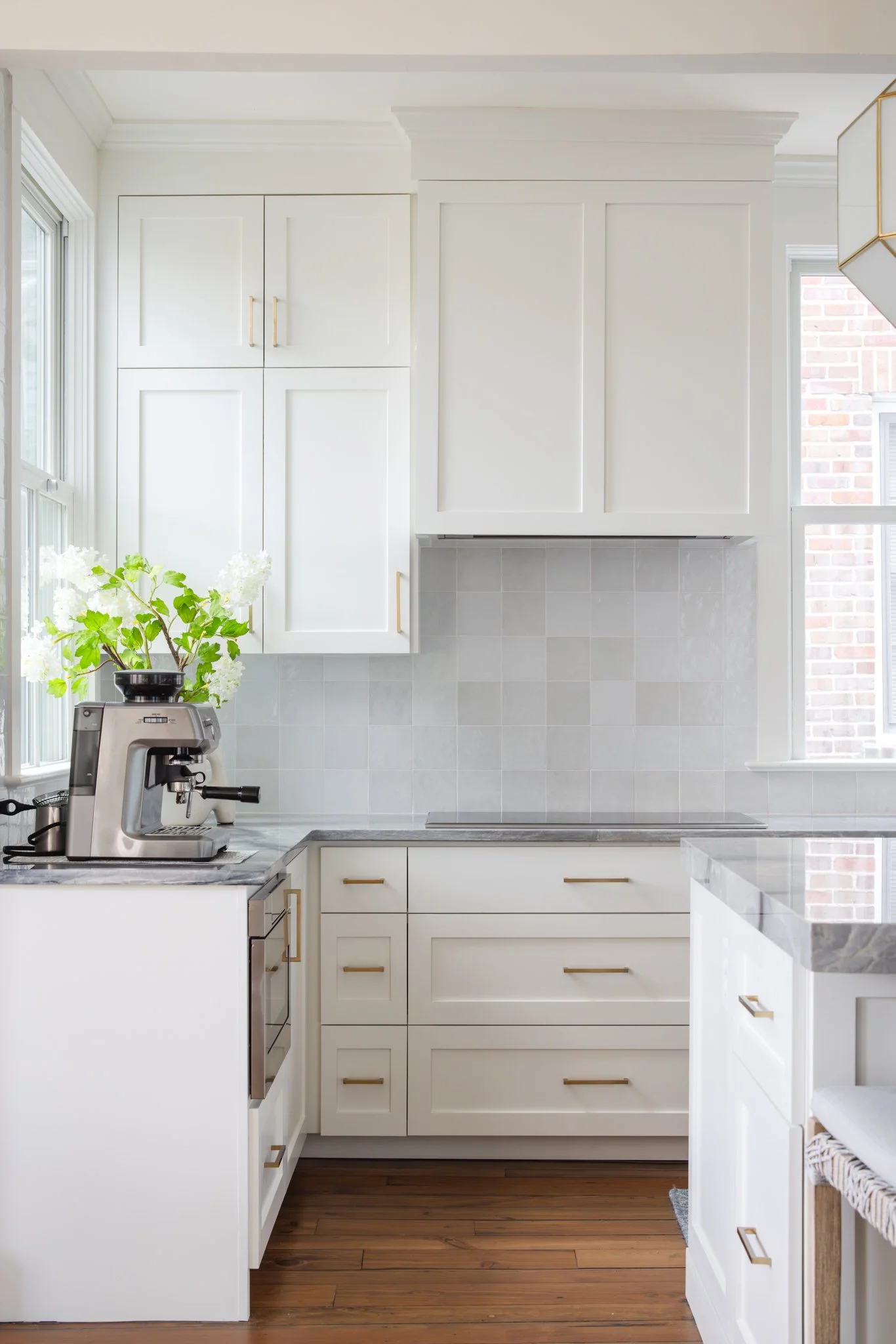 Bright white kitchen with white cabinets and gold handles, gray marble countertops, a coffee machine with a plant on top, and a window with brick wall visible outside.