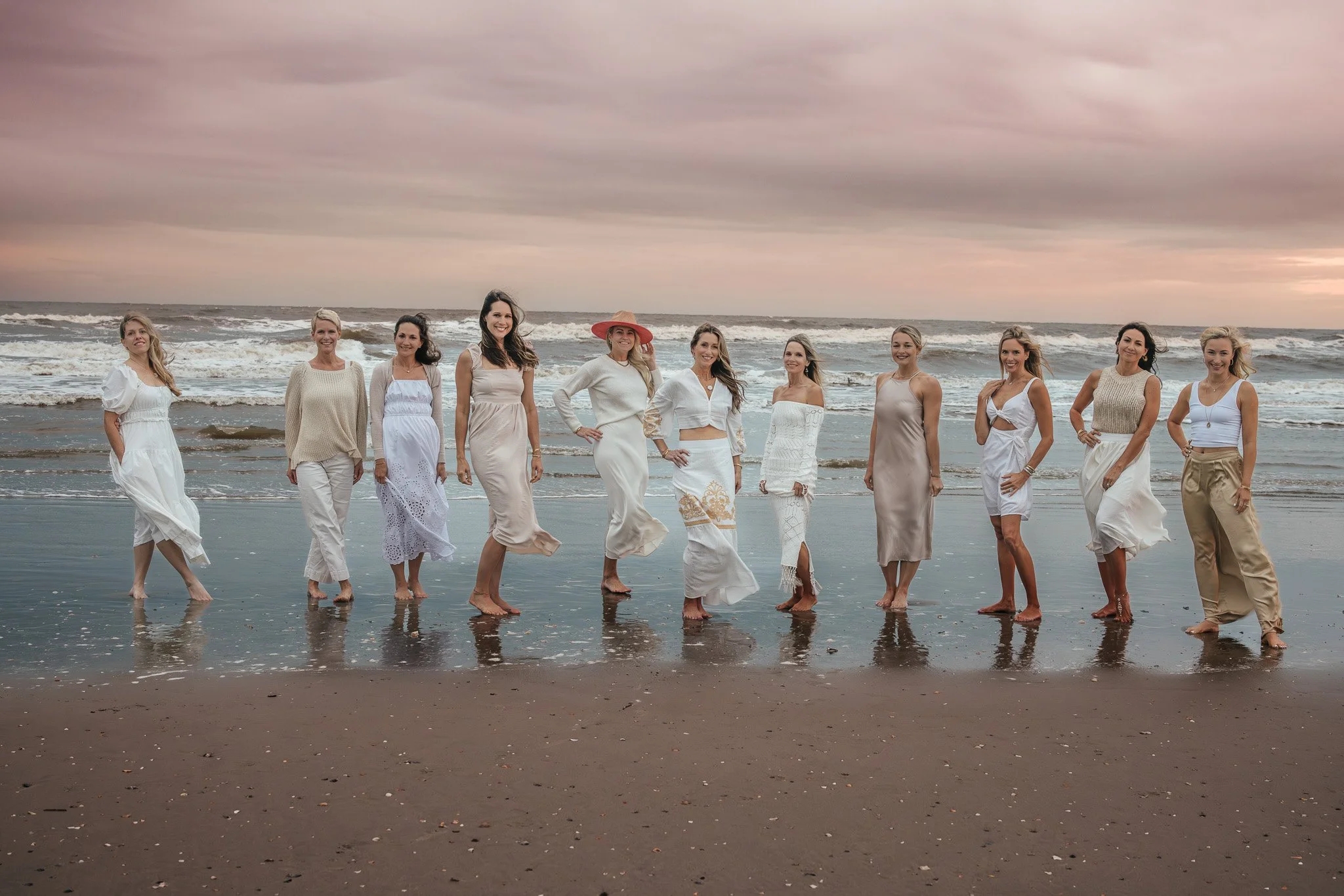 Women in white, beige, and neutral-toned dresses standing in a line at the beach with waves and a cloudy sky in the background.