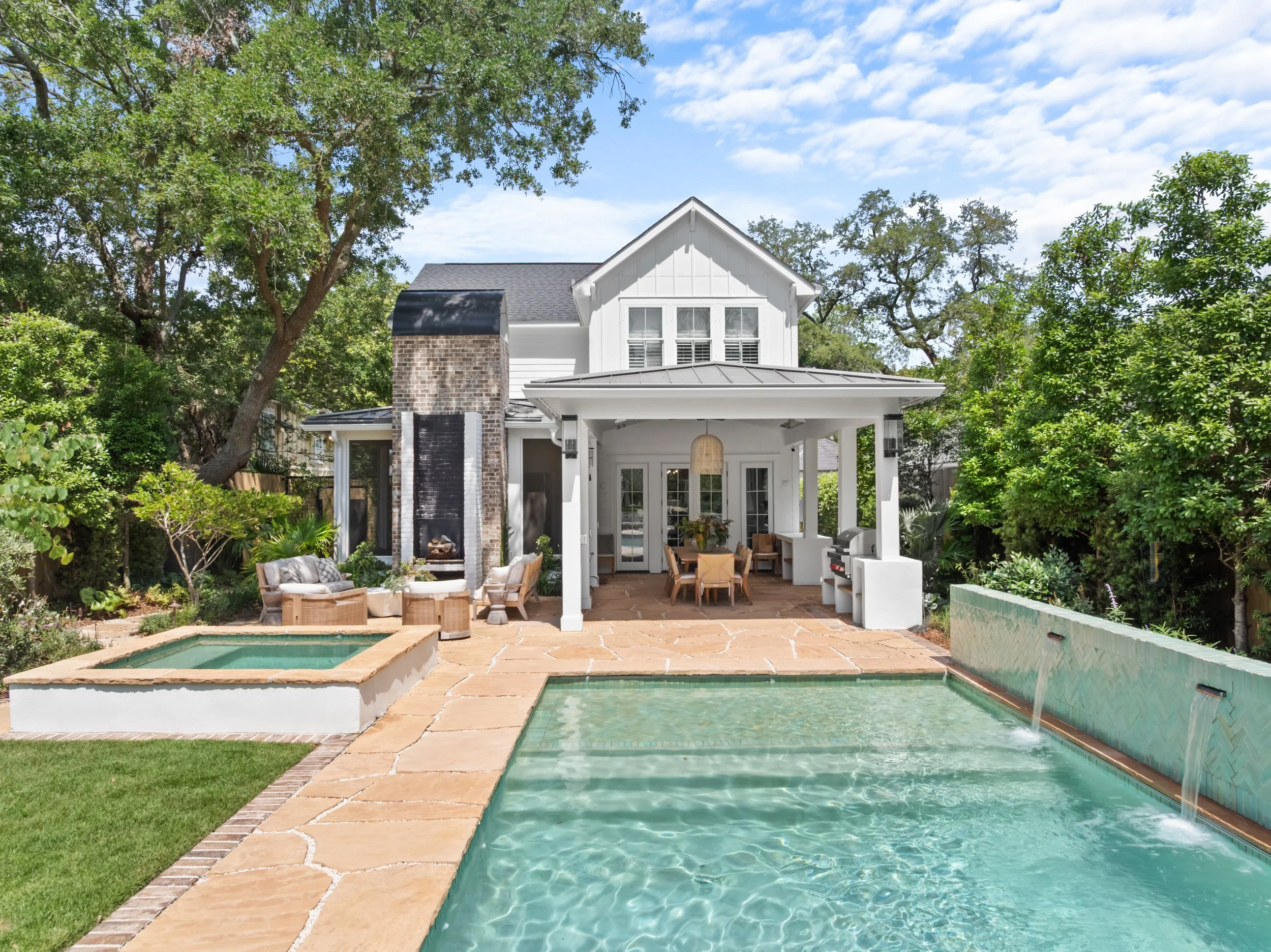 Backyard with swimming pool, hot tub, patio, outdoor fireplace, dining area, lush greenery, and a white house in the background.