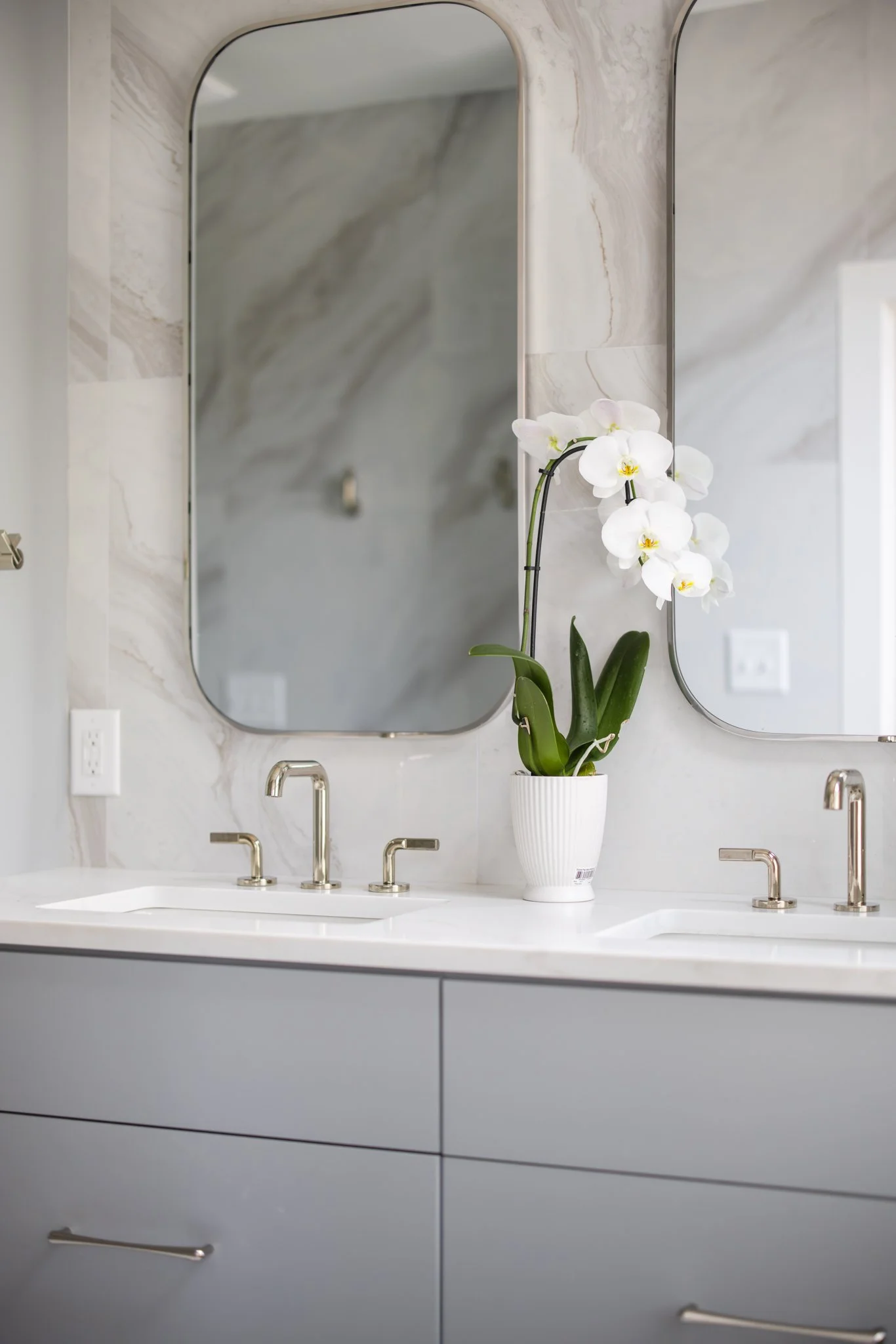 A bathroom with a marble wall, two oval mirrors, a white countertop with two sinks, and a potted white orchid plant between the mirrors.