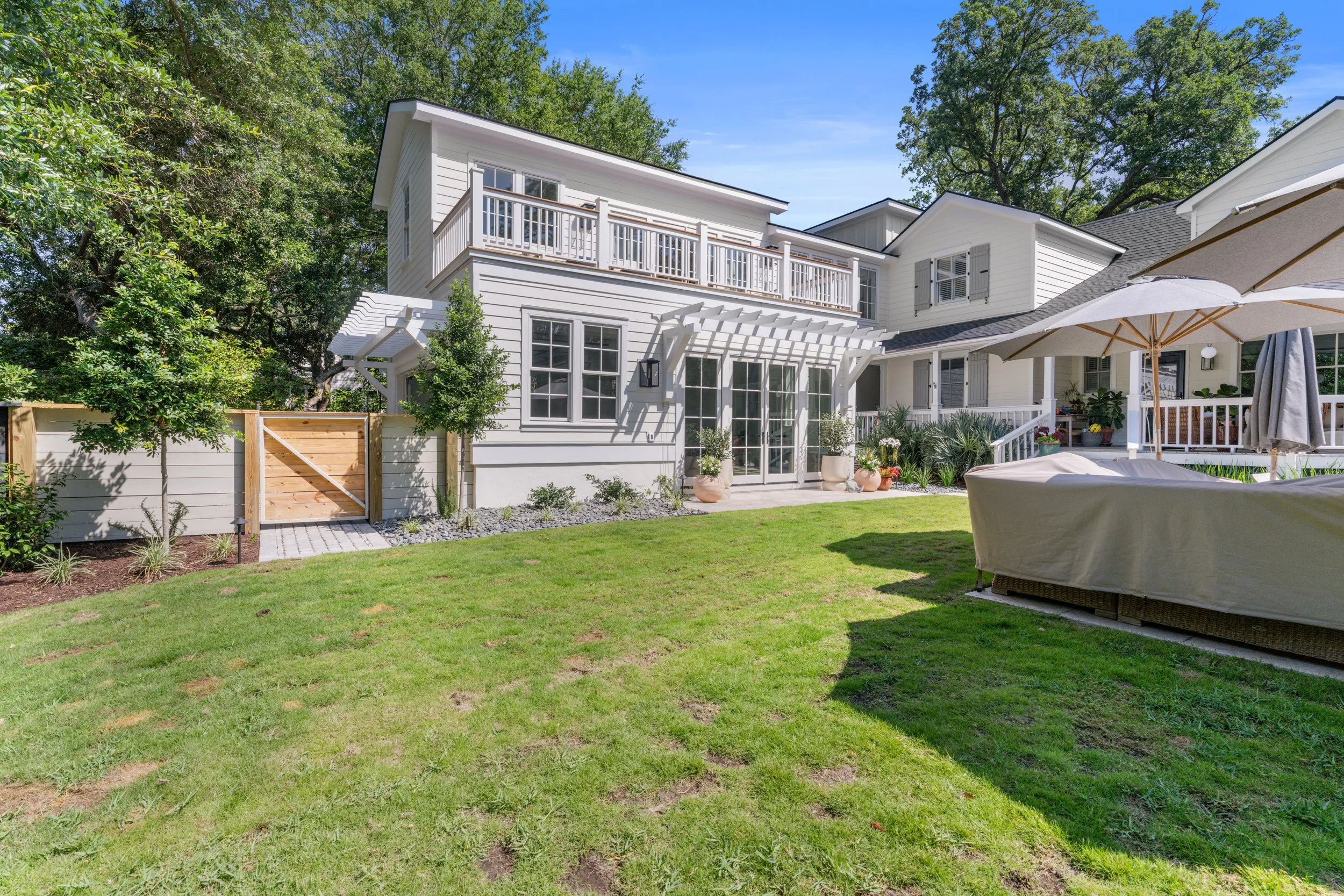 Backyard of a white house with a lawn, patio furniture, and potted plants, surrounded by trees and a wooden fence.