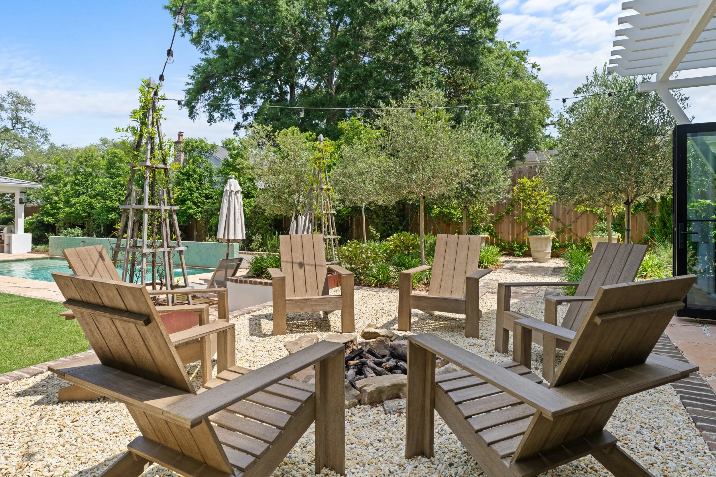 Backyard patio with six wooden Adirondack chairs arranged in a circle around a small fire pit, surrounded by greenery, trees, and string lights, with a pool and outdoor furniture in the background.