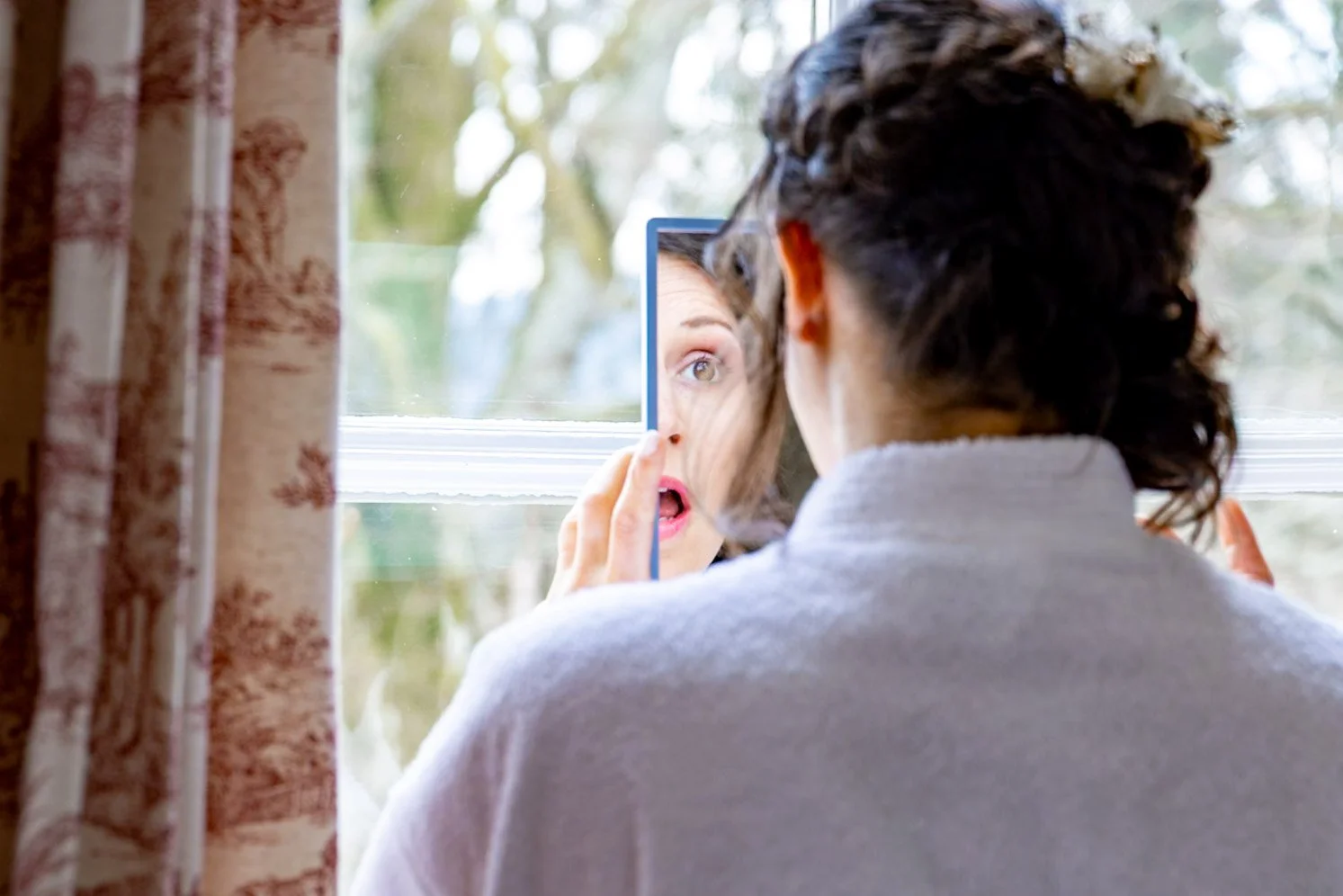 Femme regardant son reflet dans un miroir de poche, vue depuis derrière, à côté d'une fenêtre avec rideaux en tissu. Le fond montre des arbres outside.