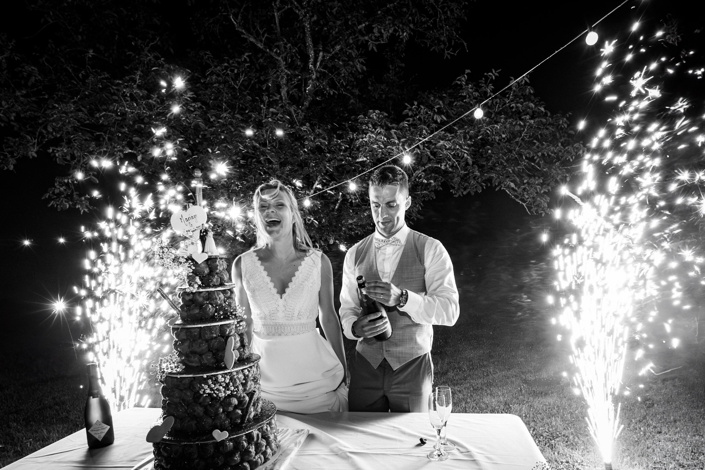 Un couple souriant lors d'une célébration de mariage à l'extérieur, avec un gâteau de mariage à plusieurs niveaux décoré de fruits, des bouteilles de champagne, des flambeaux et des feux d'artifice en arrière-plan.