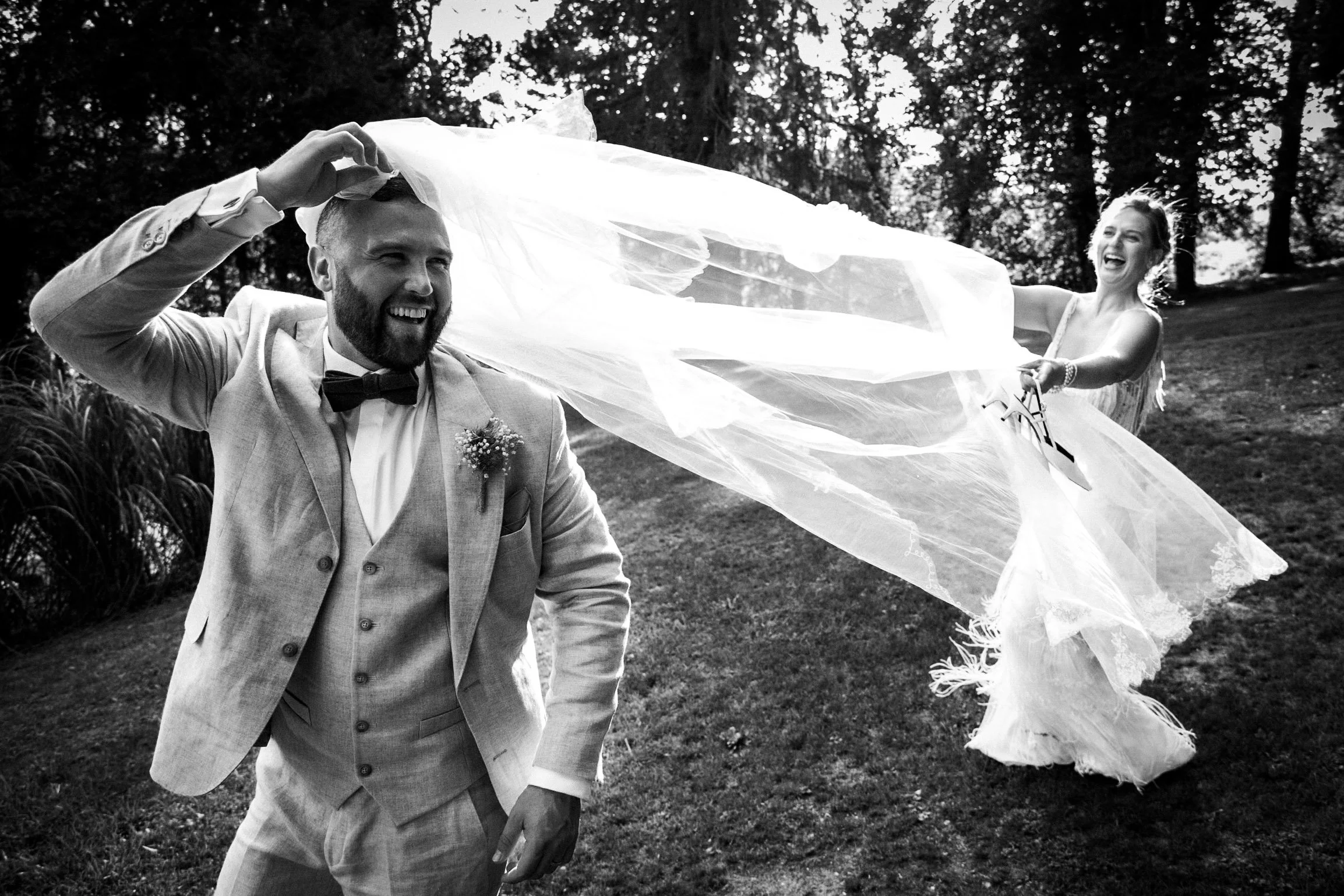 Un homme et une femme en mariage dans un cadre extérieur, l'homme souriant et la femme s'amusant avec un voile, la femme en robe de mariage et l'homme en costume avec nœud papillon.