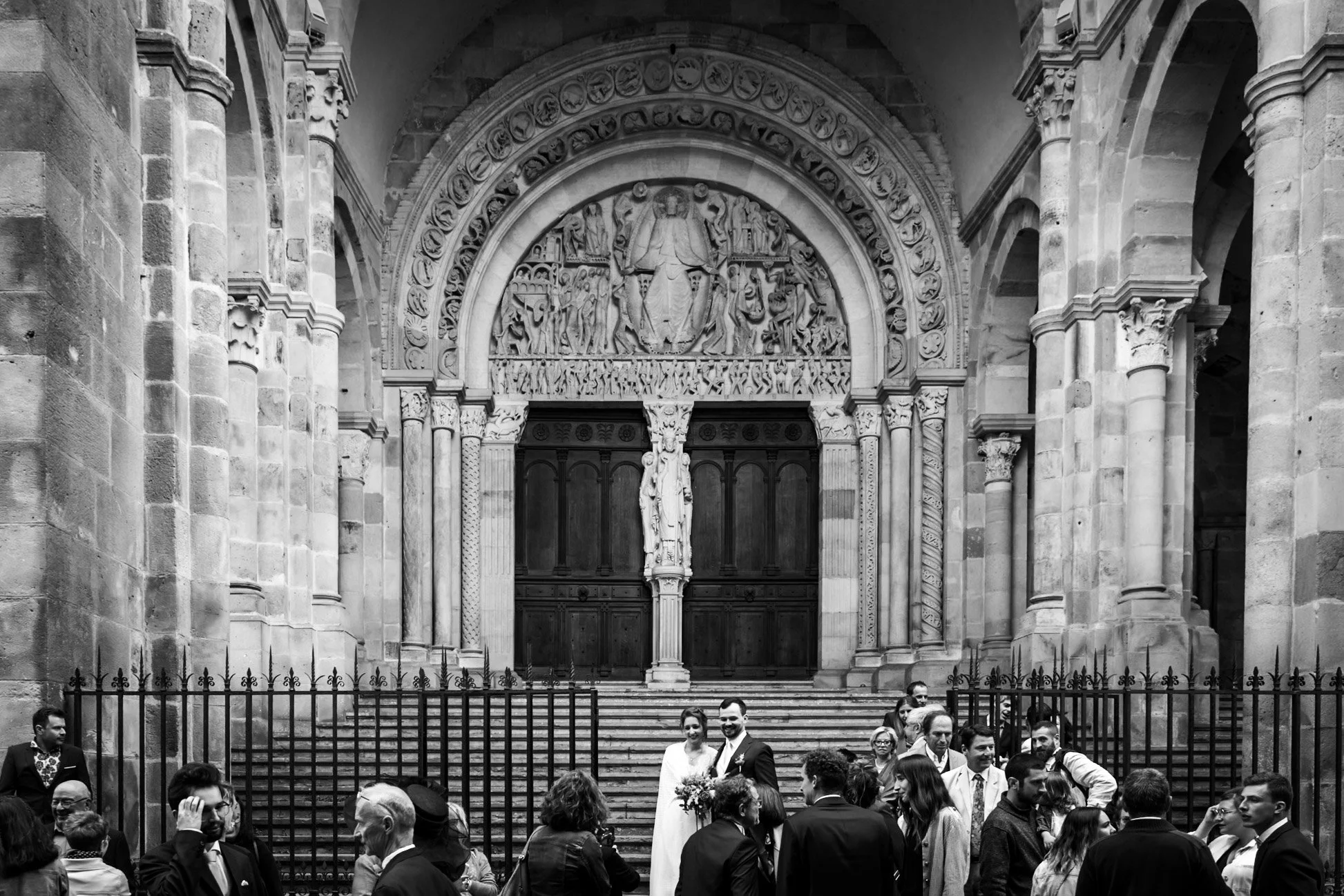 Photo en noir et blanc d'un groupe de personnes devant l'entrée d'une église ou cathédrale, avec une sculpture au-dessus des portes et des escaliers. Un couple en vêtements de mariage se trouve au centre, entouré d'invités.