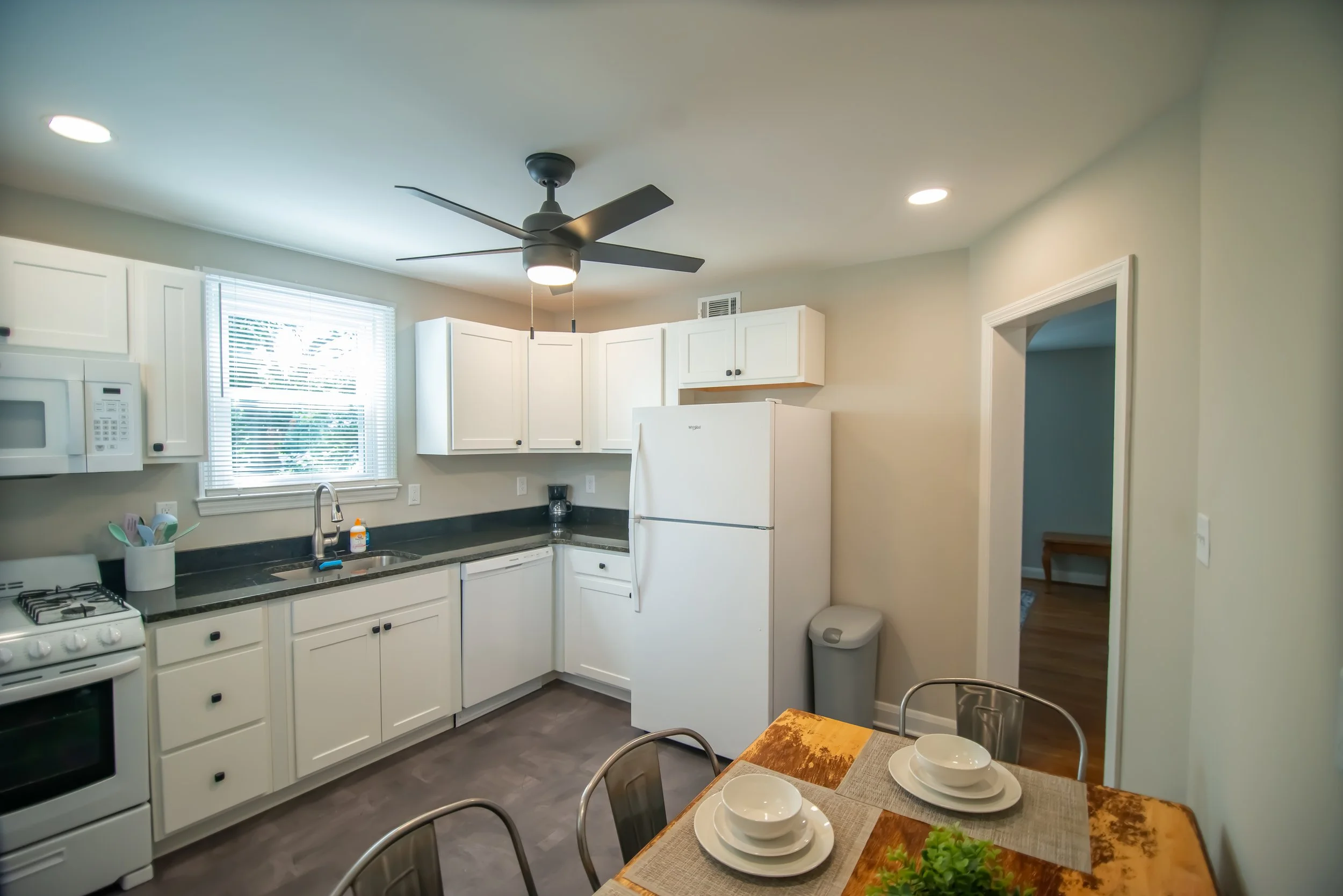 Kitchen with white cabinets, microwave, refrigerator, stove, window, ceiling fan, dining table with placemats, bowls, and green salad.