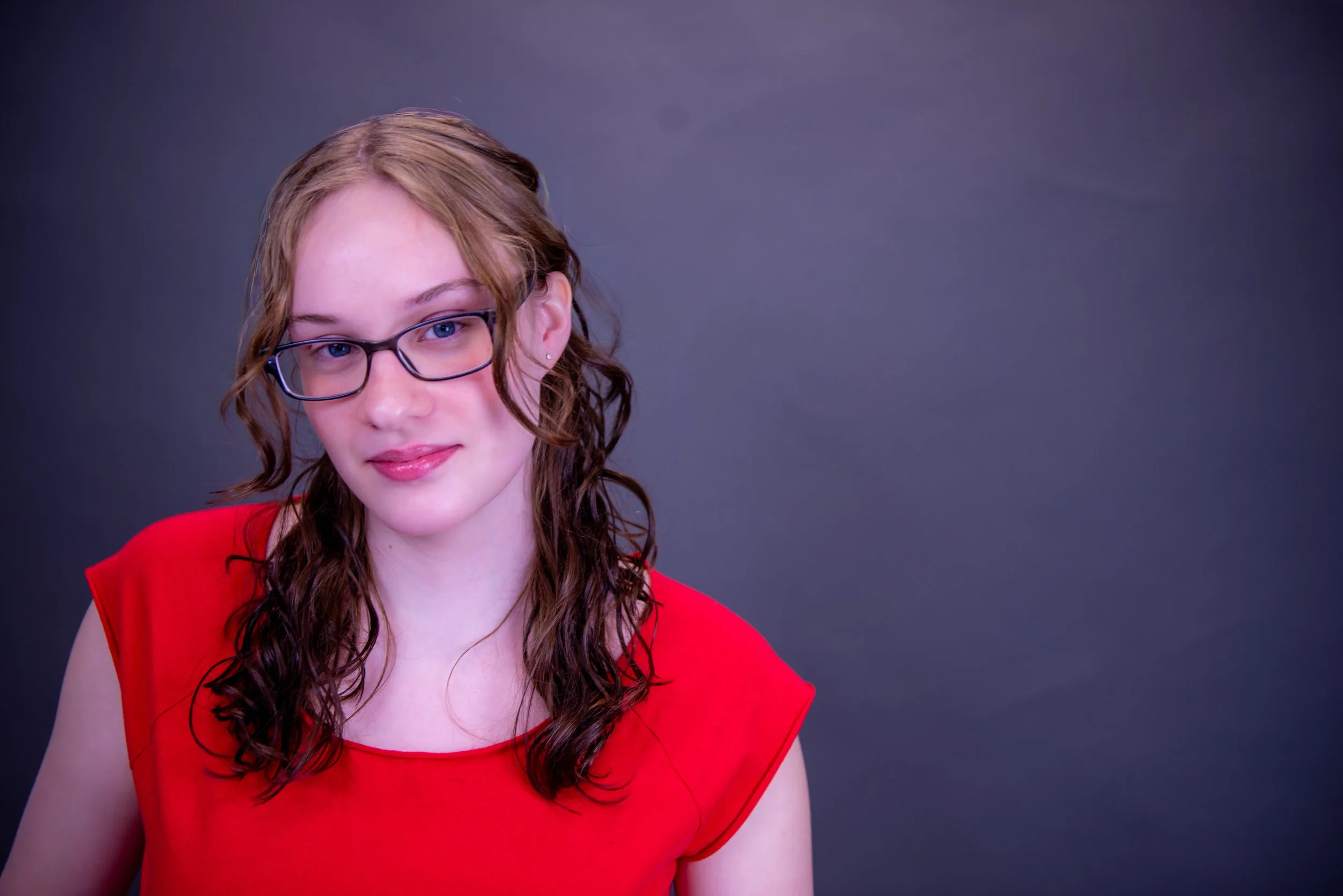 A young woman with curly brown hair, wearing glasses and a red sleeveless top, is looking at the camera against a plain dark background.