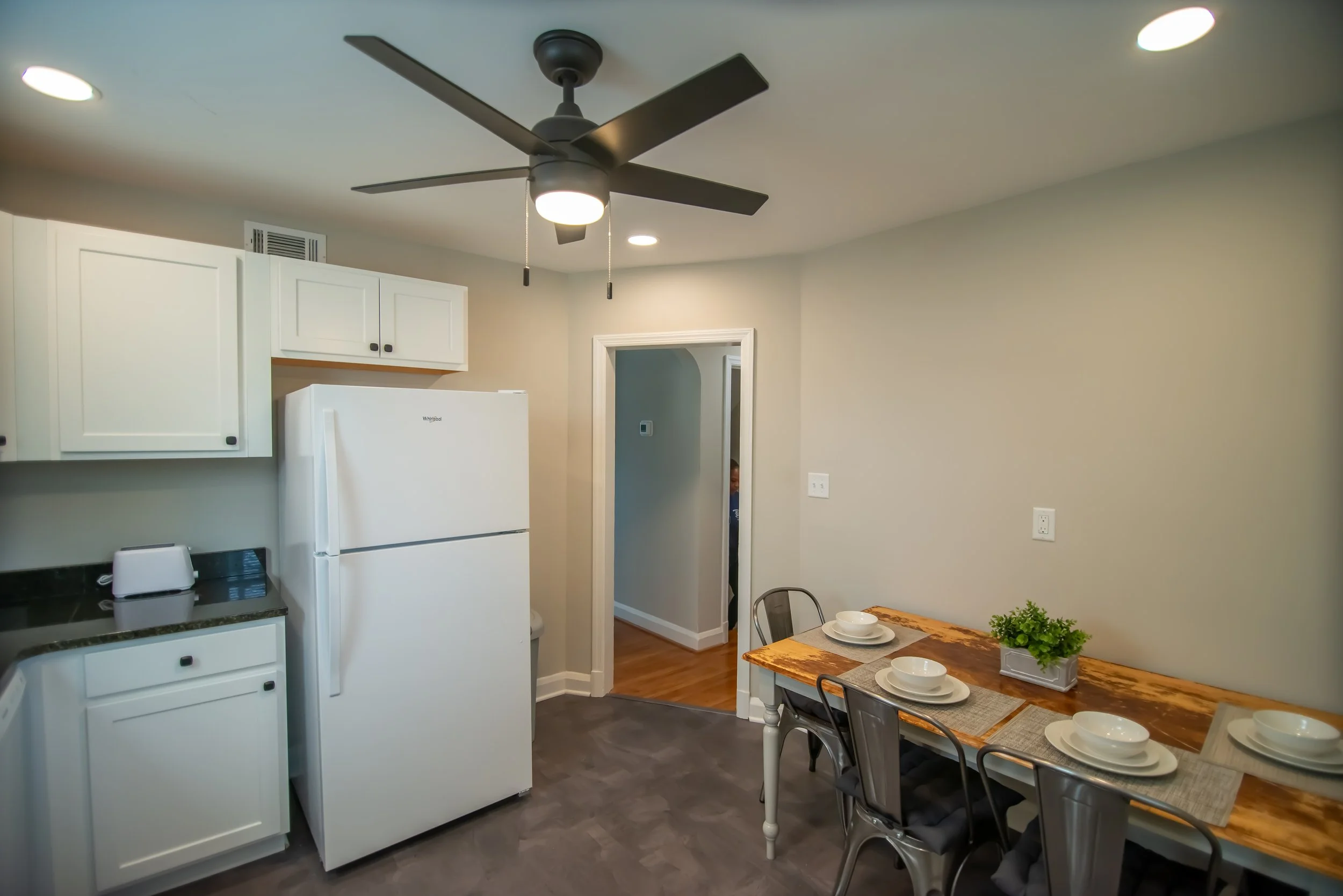 Kitchen with white cabinets, a white refrigerator, a ceiling fan, and a dining table set with white plates and bowls, along with a small green plant.