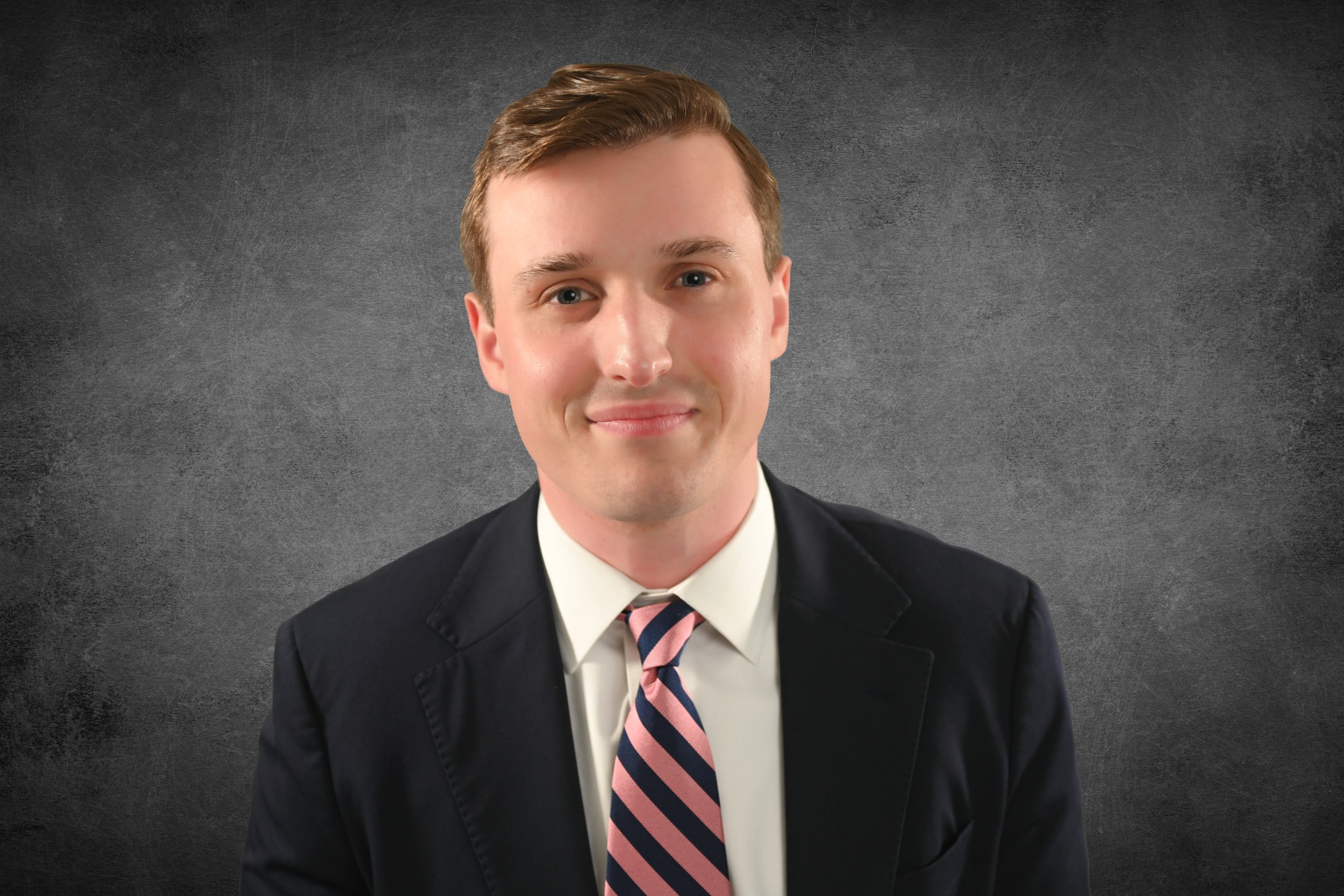 A young man with short light brown hair, wearing a black suit, white shirt, and a striped pink and navy tie, smiling slightly against a dark, textured background.