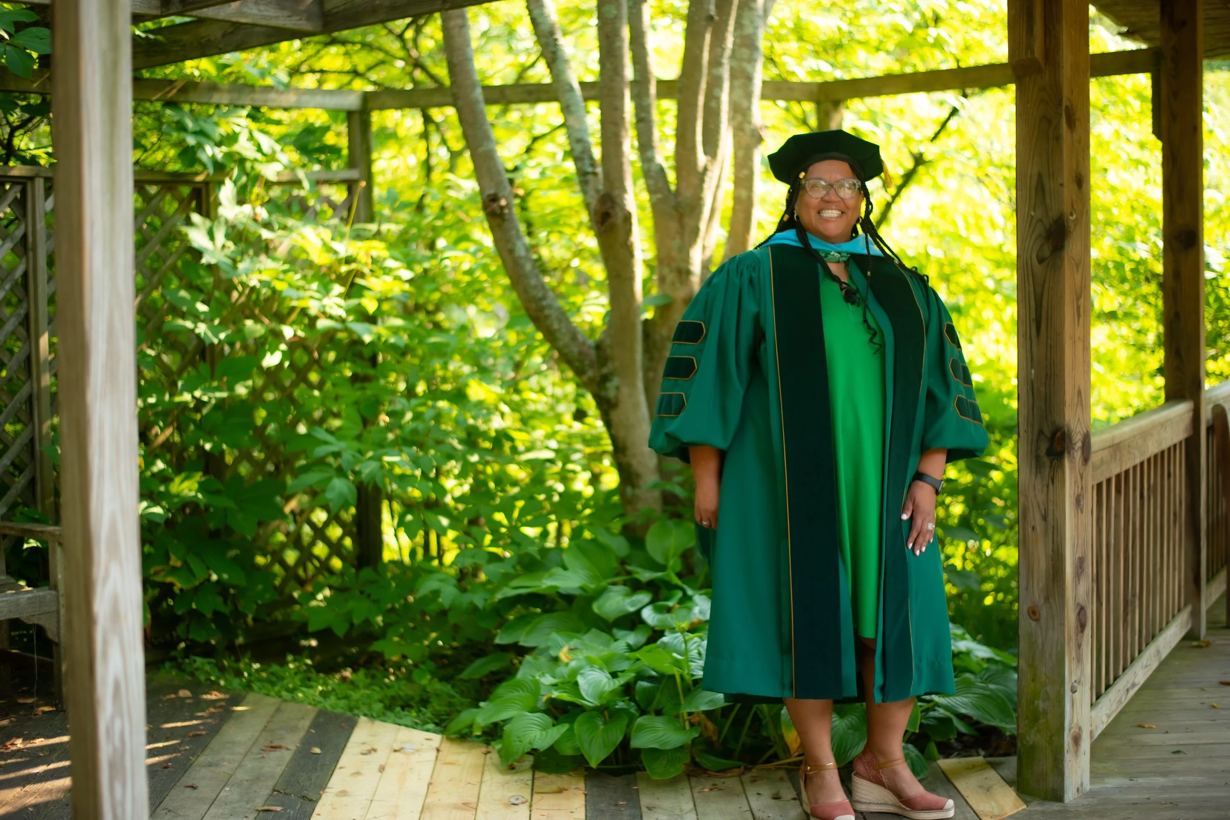 Woman in green academic gown and cap standing on wooden bridge in a lush green outdoor setting during graduation ceremony.
