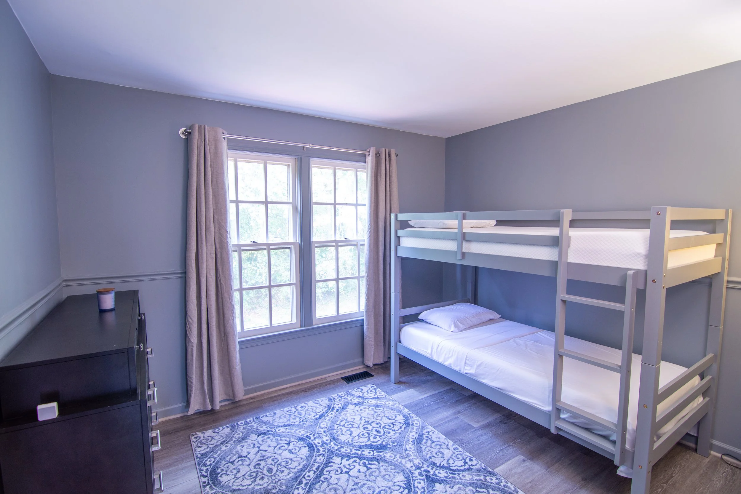 A bedroom with light blue walls, a window with white trim and beige curtains, a black dresser with candles on top, a white bunk bed with pillows and bedding, a patterned rug, and hardwood flooring.