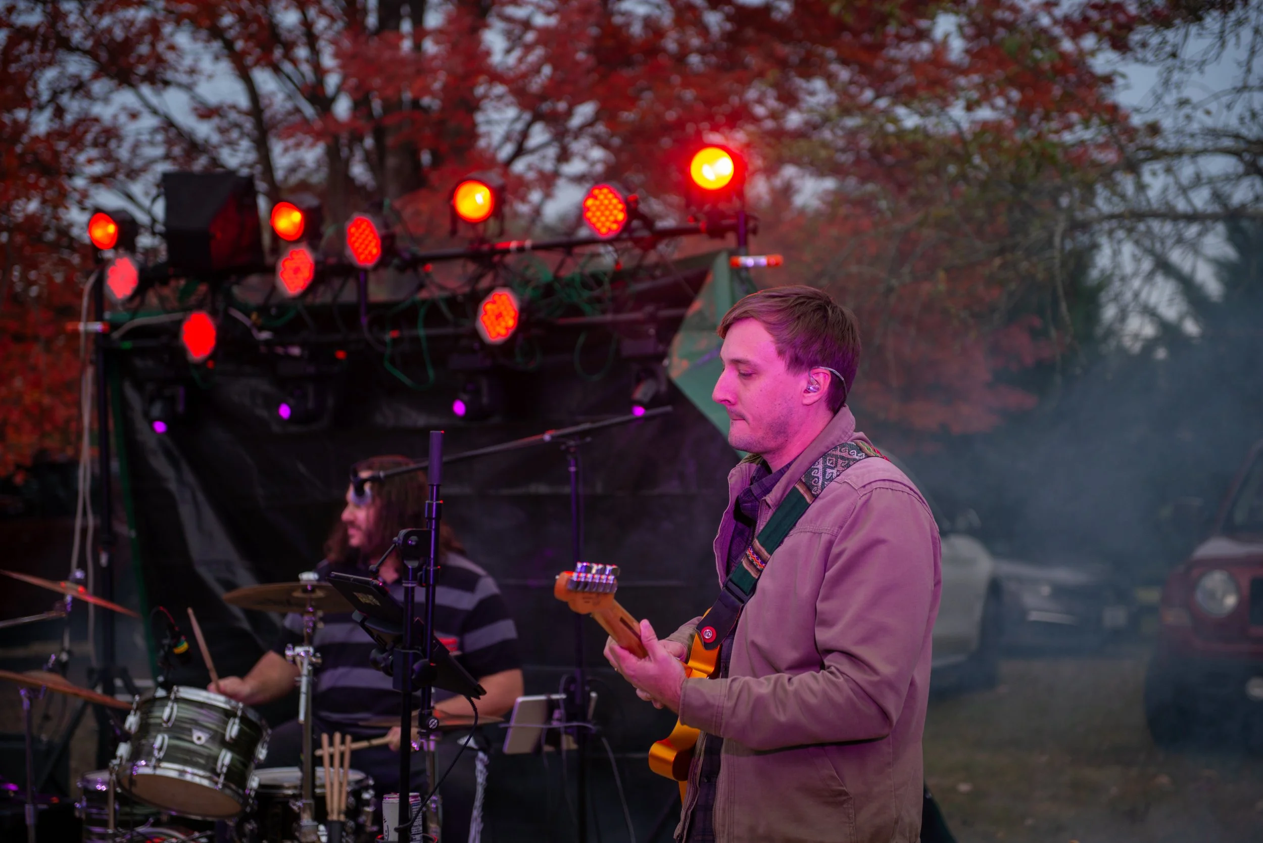 A musician playing an electric guitar during an outdoor performance at dusk, with a tree with red leaves and stage lights in the background.