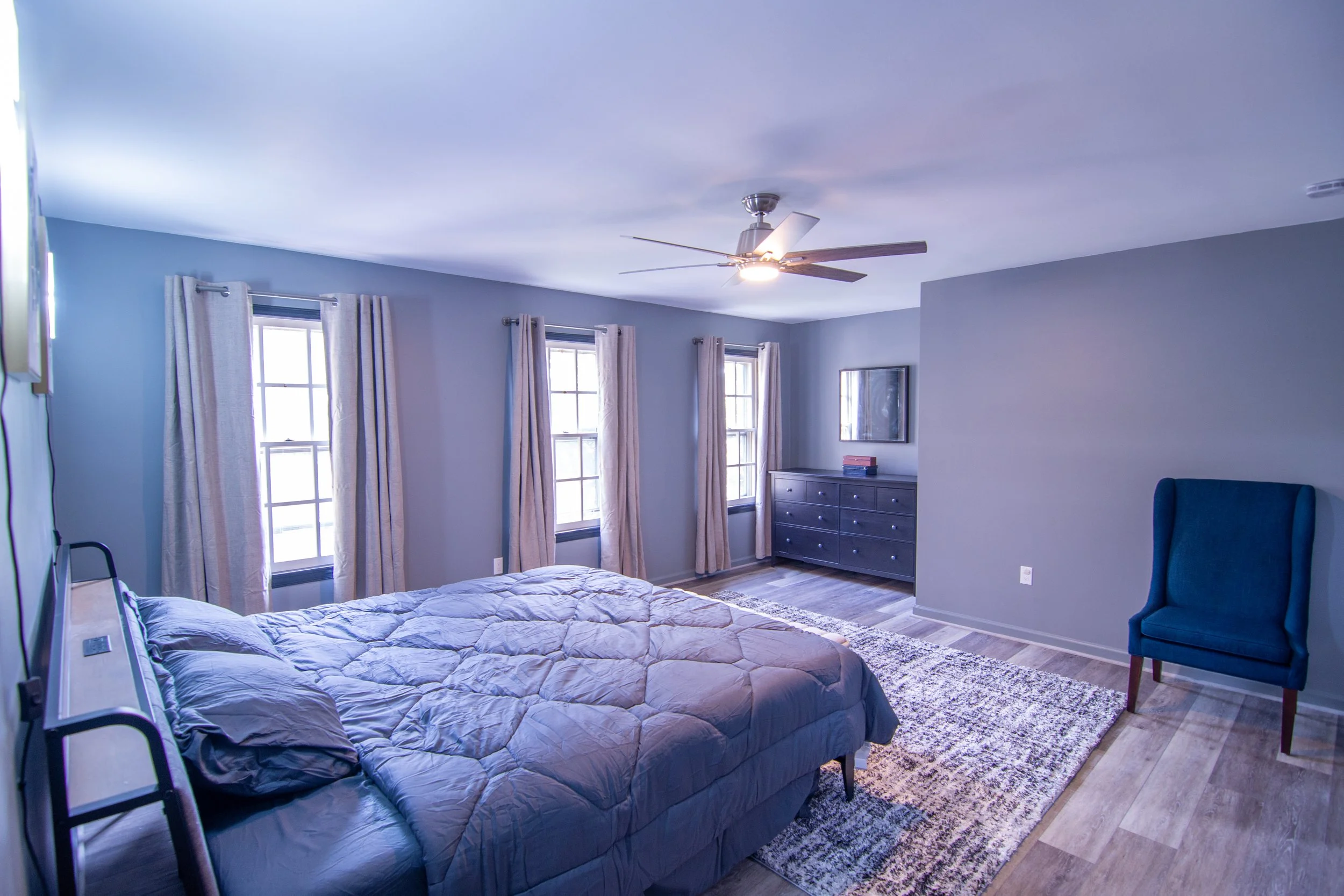 Bedroom with light blue walls, three windows with light curtains, ceiling fan, dresser with books, bed with light-colored bedding, blue armchair, and area rug.