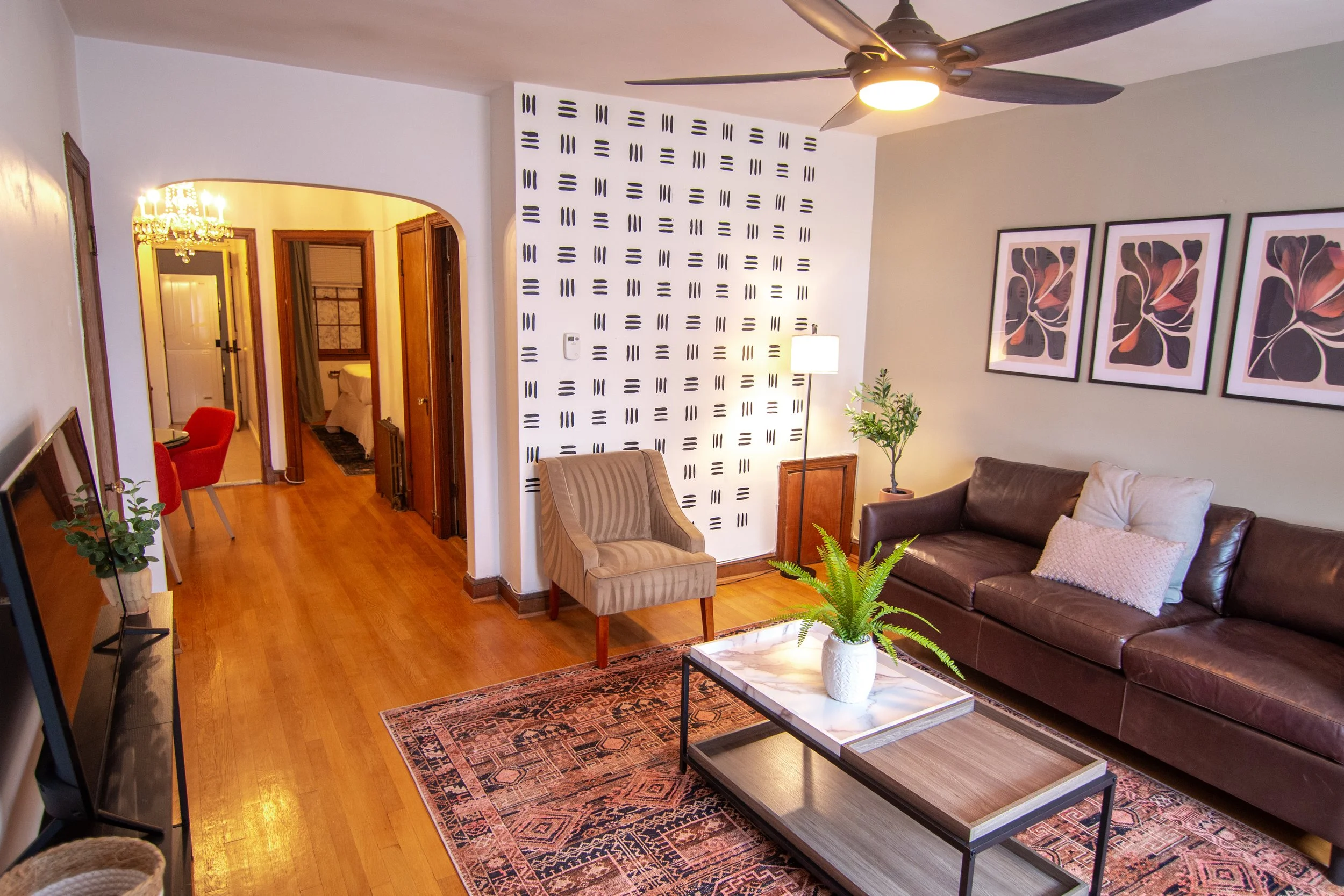 Living room with wooden floors, a dark brown leather couch with pillows, a marble coffee table, a small potted plant, and framed abstract art on light-colored walls. An archway leads to a bedroom, and a patterned rug is partially visible.