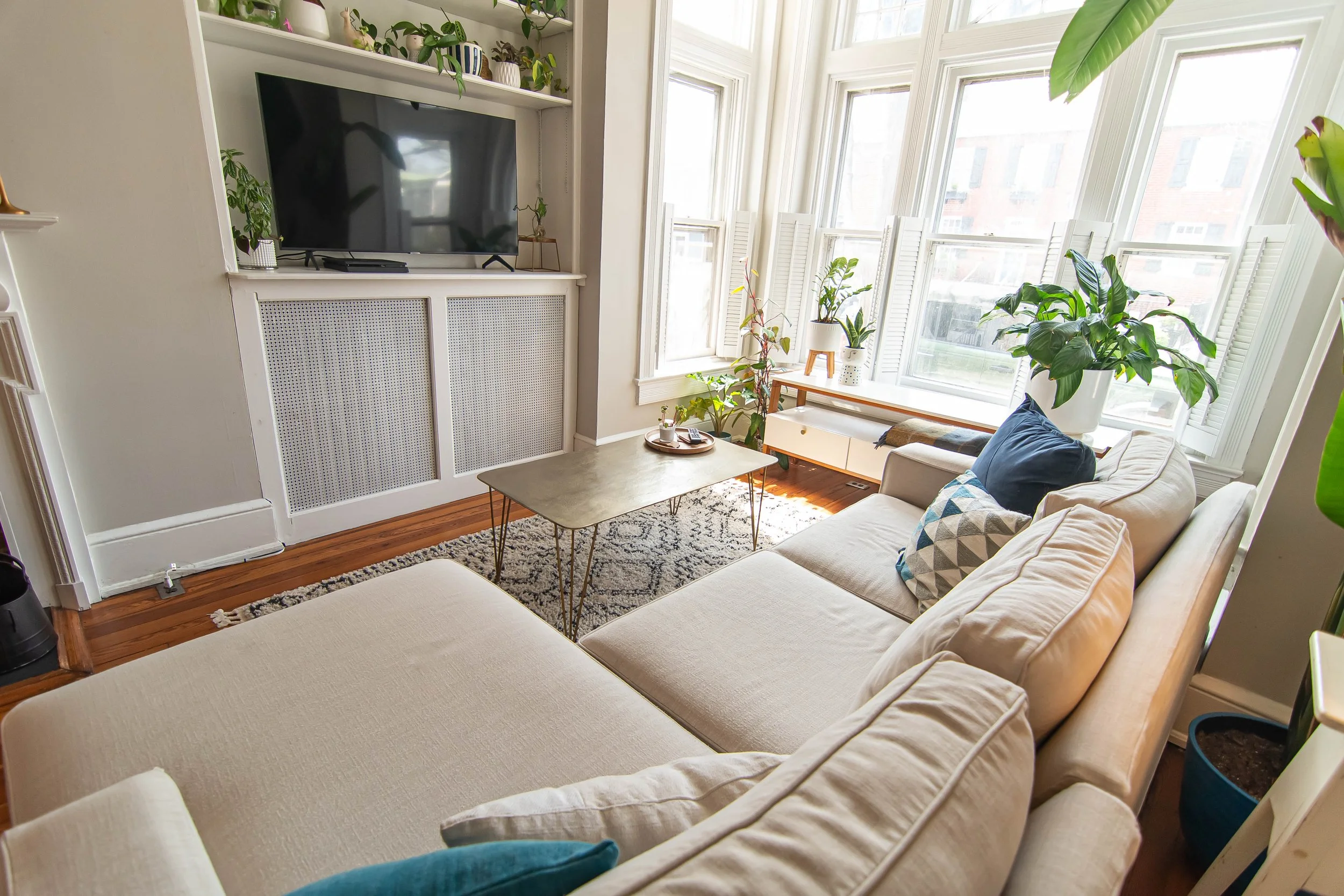 A cozy living room with large bay windows letting in natural light. The room features a beige sectional sofa with patterned and solid pillows, a rectangular coffee table, and numerous green potted plants. A white media cabinet with a flat-screen TV a