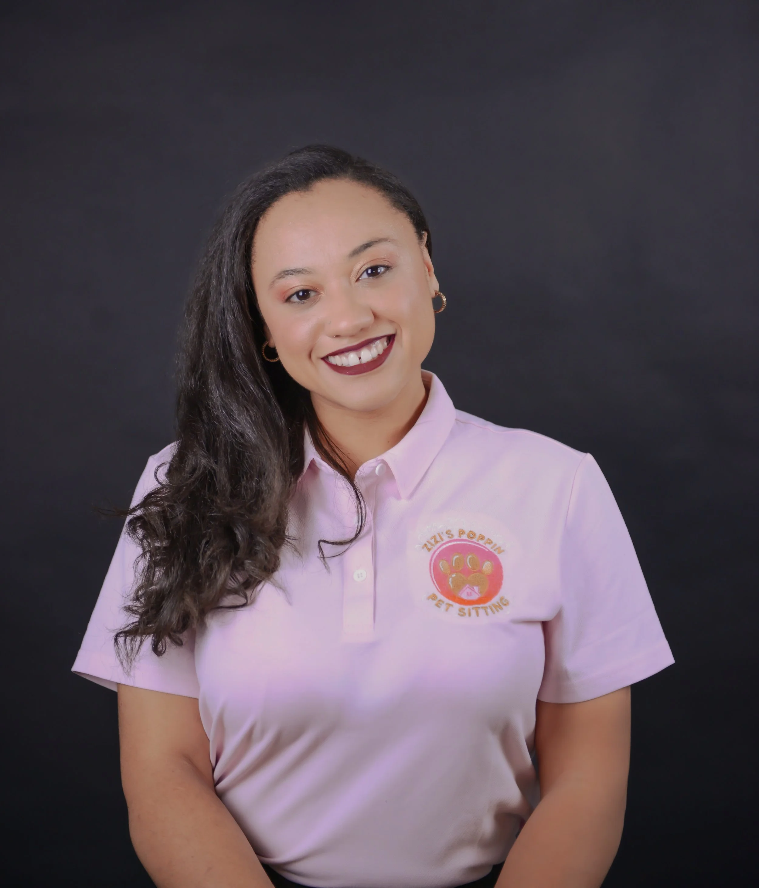 A woman with long, curly dark hair smiling against a black background, wearing a light pink polo shirt with a logo that reads "Zizi's Poppin Pet Sitting."