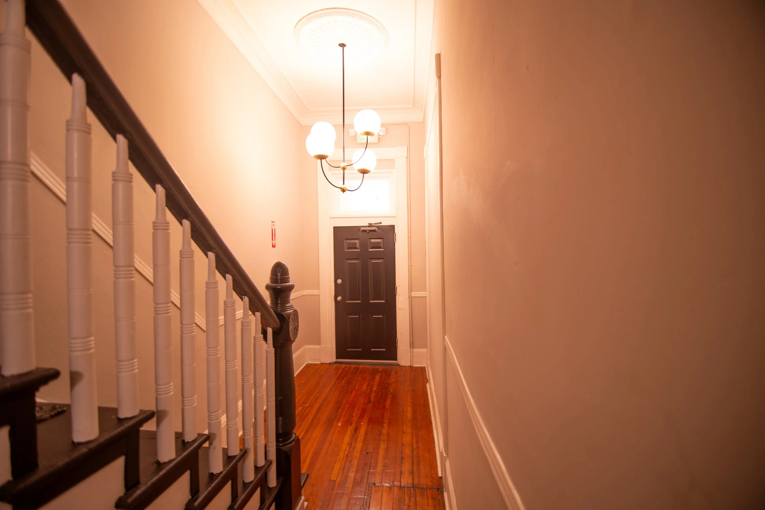 Indoor hallway with wooden flooring, a staircase with white spindles on the left, a black front door at the end, and a ceiling light fixture with multiple white globes.