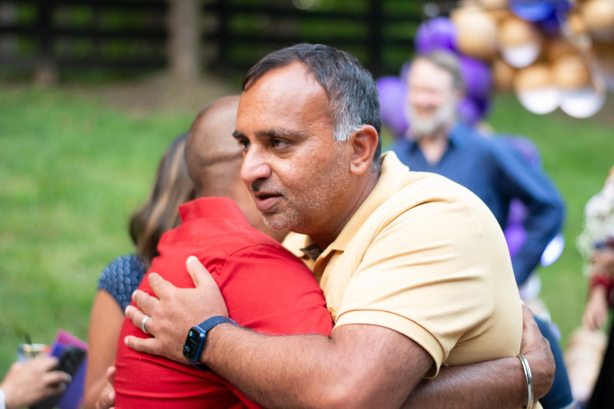 A man and a woman are hugging outdoors, with other people and balloons in the background.