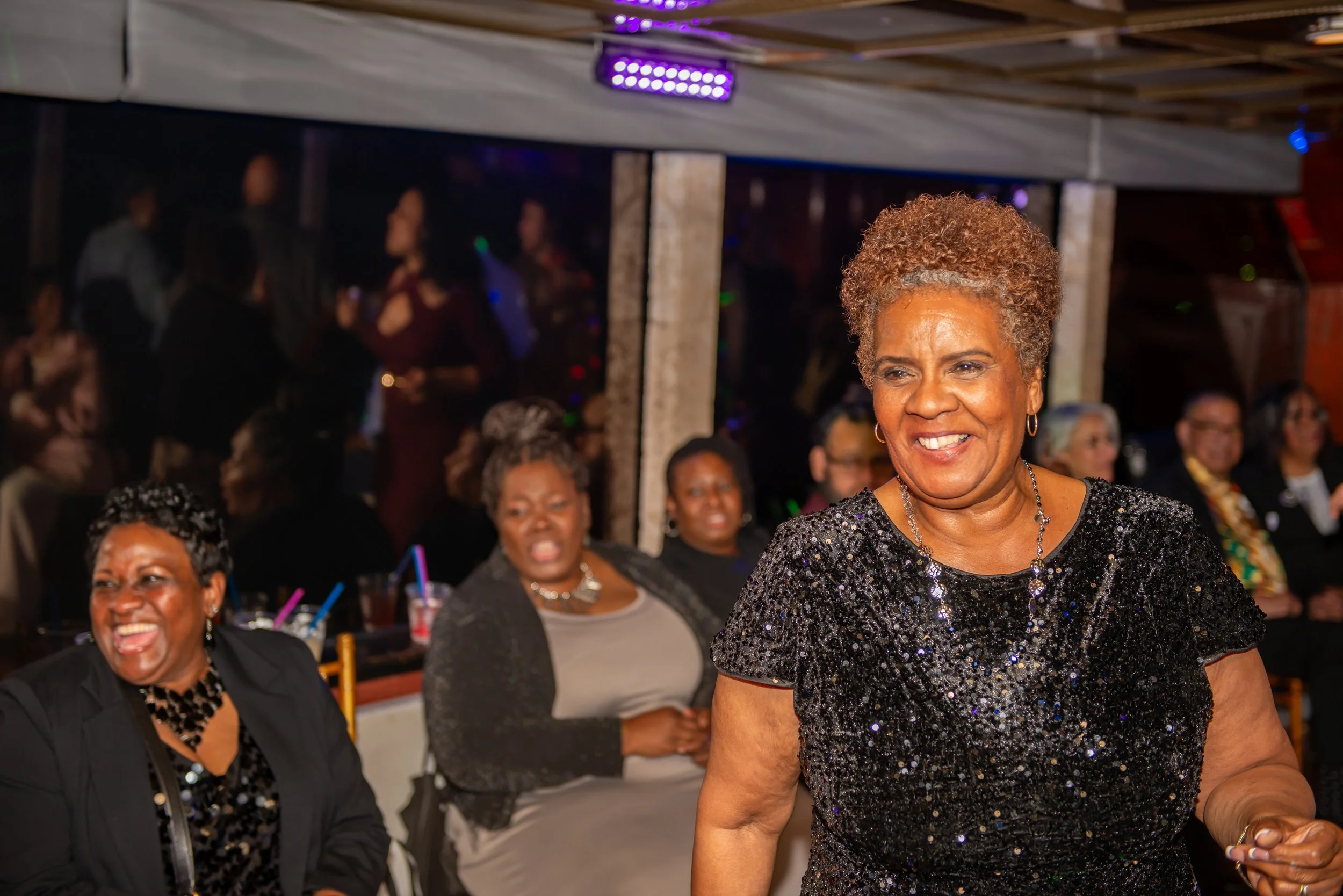 An older woman with curly, short, reddish-brown hair wearing a black, sequined dress, smiling and standing at a social gathering with several predominantly African American women in the background.
