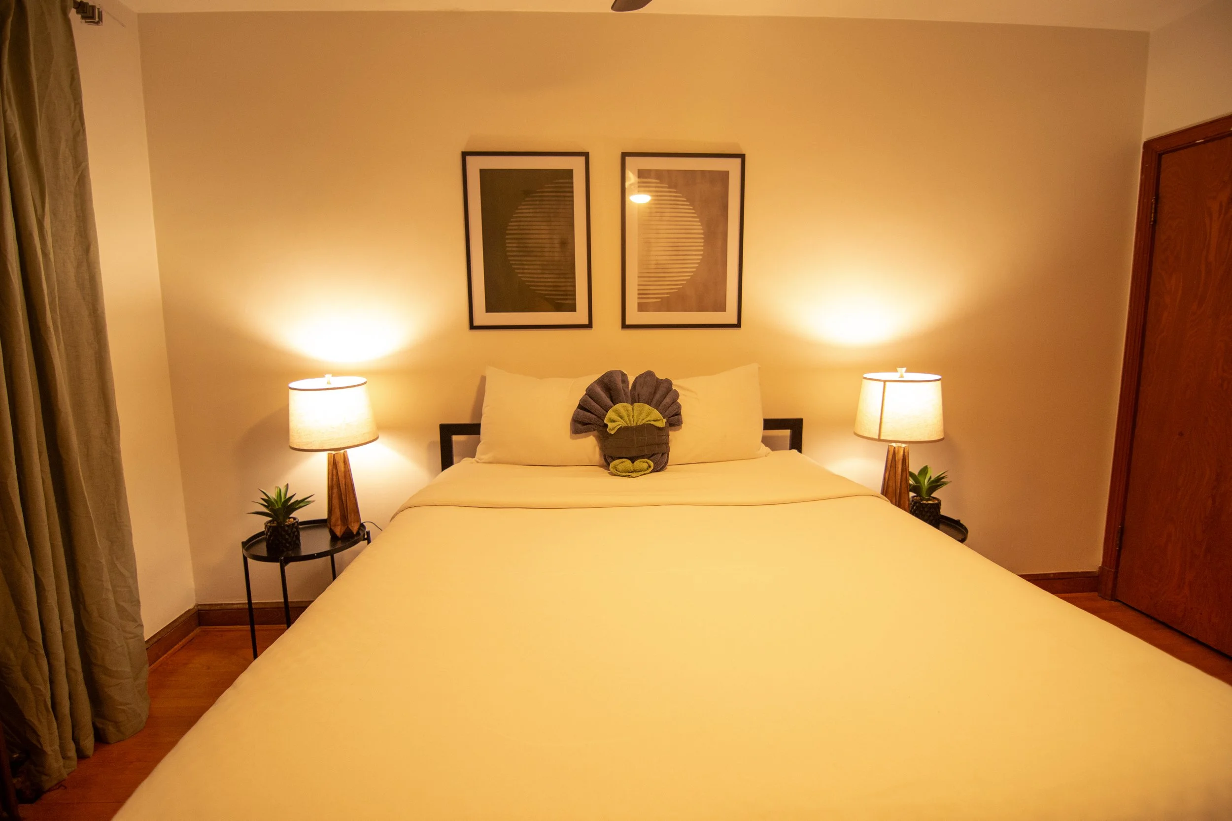 A cozy bedroom with a neatly made bed, two nightstands with lamps, and framed artwork above the headboard. The room features warm lighting and wooden accents.