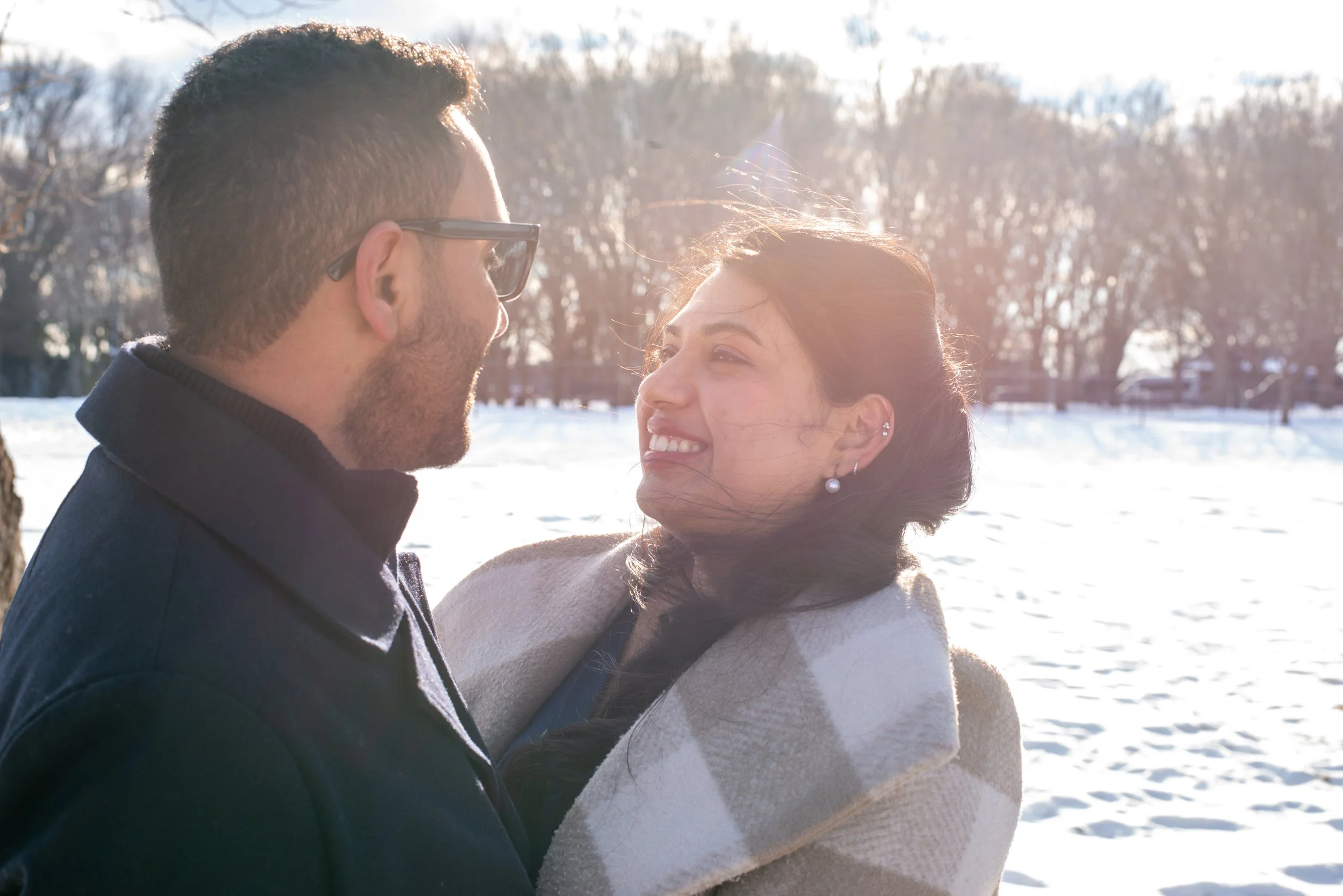 A couple smiling and looking at each other outdoors in a snowy landscape with bare trees and sunlight in the background.