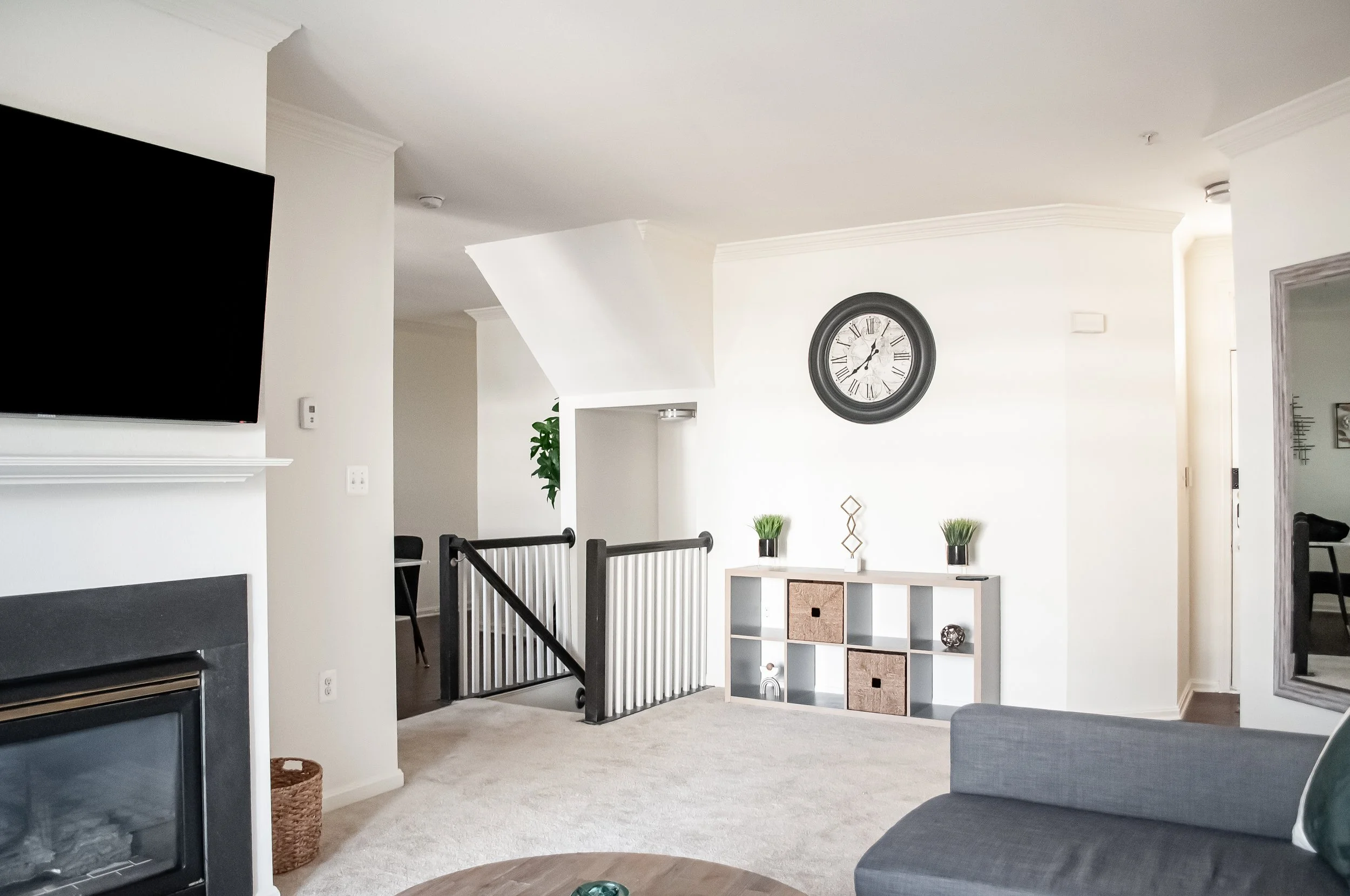Living room with white walls, black-framed wall clock, a white shelving unit with decorative items, a gray armchair, and a TV in the corner. There is a staircase with black railings and a plant in the background.