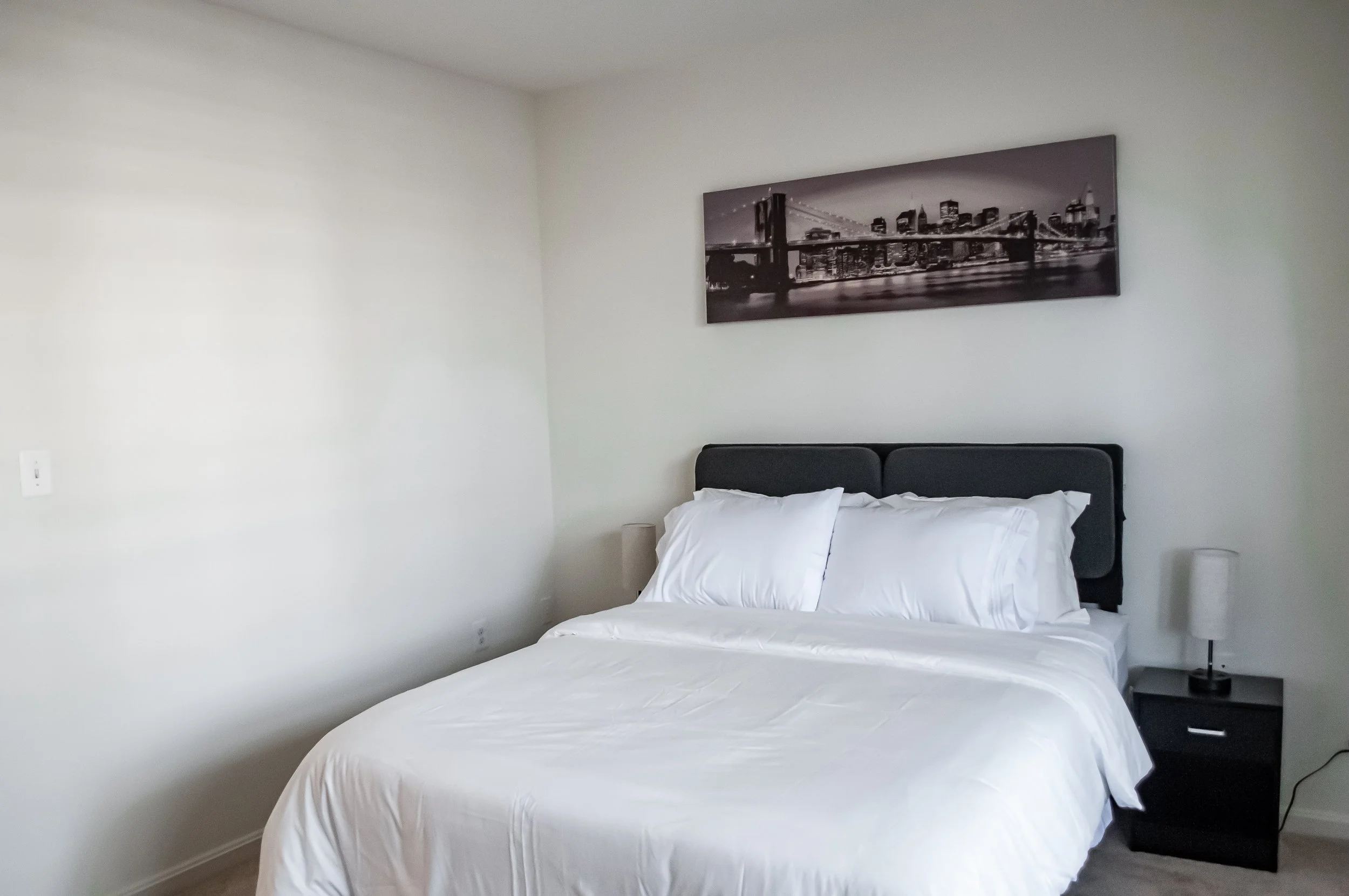A bedroom with a bed covered in white sheets and pillows, a black headboard, side table with a black lamp, and a black and white cityscape photograph on the white wall.