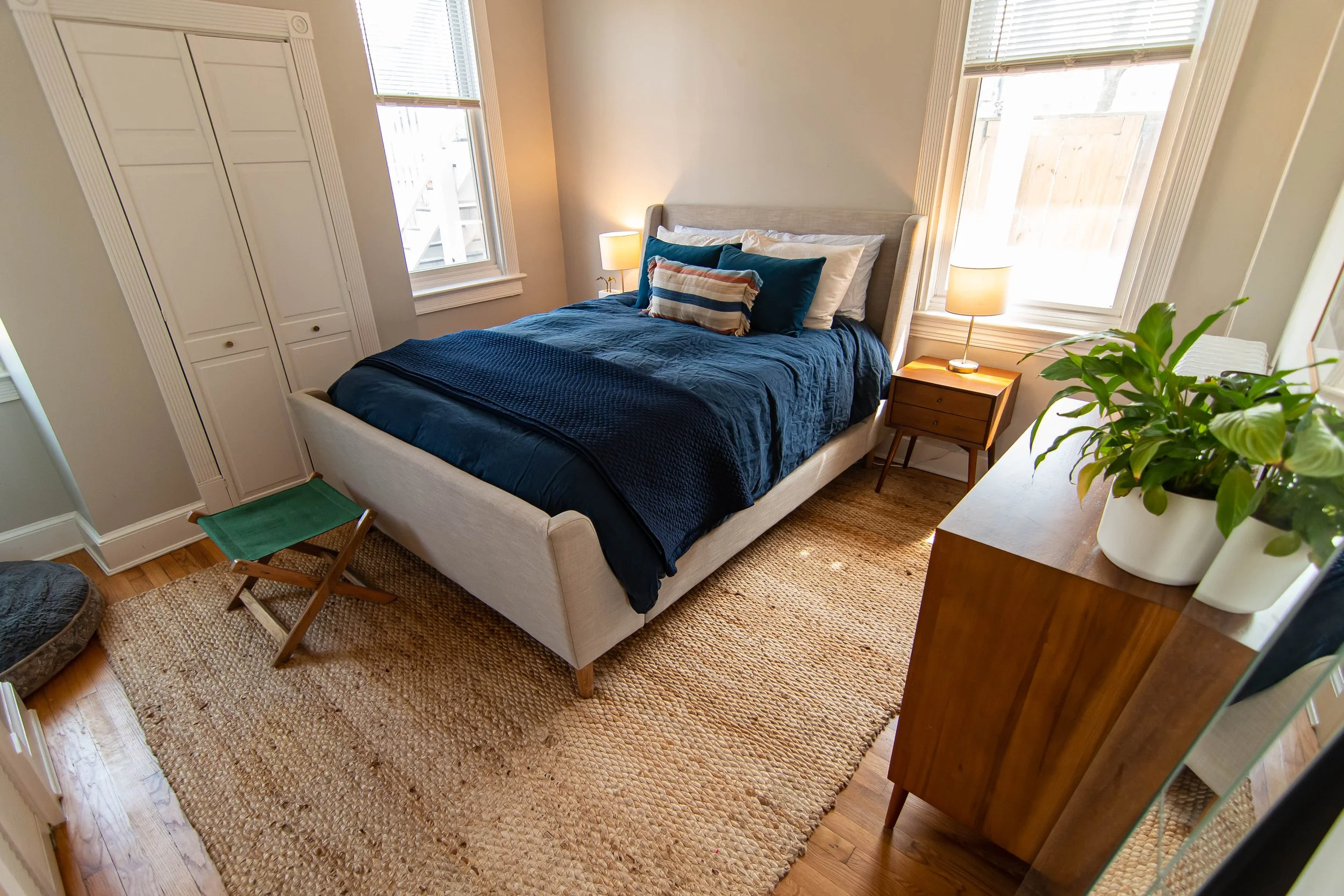 A cozy bedroom with a bed covered in a dark blue quilt and several pillows, a nightstand with a lamp, windows with blinds, a wooden cabinet, a potted plant, and a woven rug on hardwood floor.