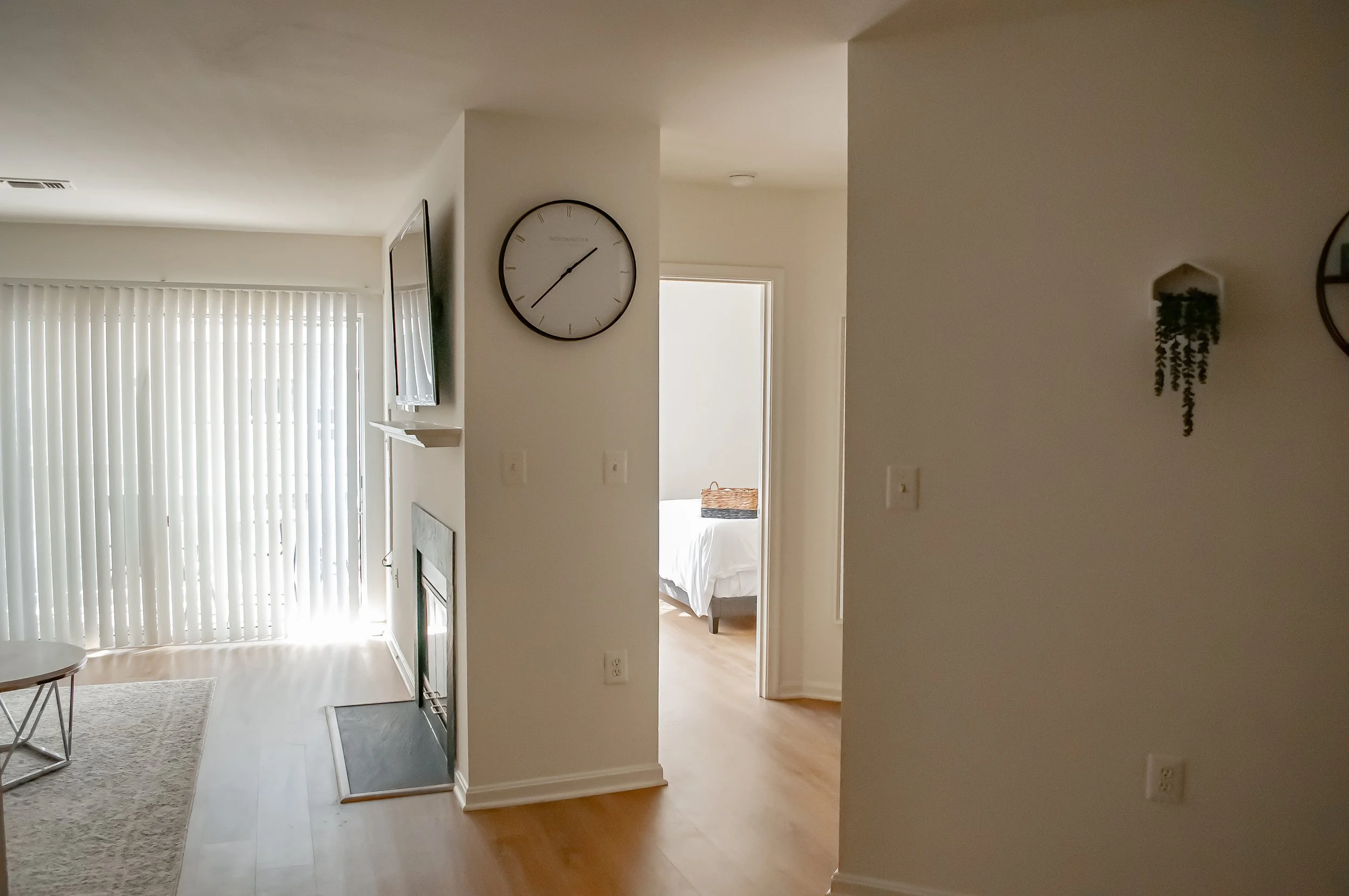 Interior view of a home with a living area and a bedroom visible through an open doorway, featuring a large wall clock, sliding glass door with vertical blinds, and minimal decor.
