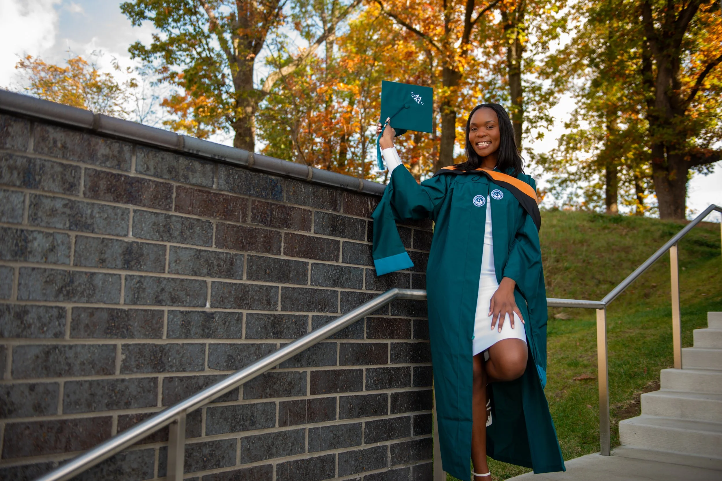 A young woman in a green graduation gown and cap stands outdoors on a staircase, smiling and holding her cap in the air. She is dressed in a white dress underneath the gown, and autumn trees are in the background.