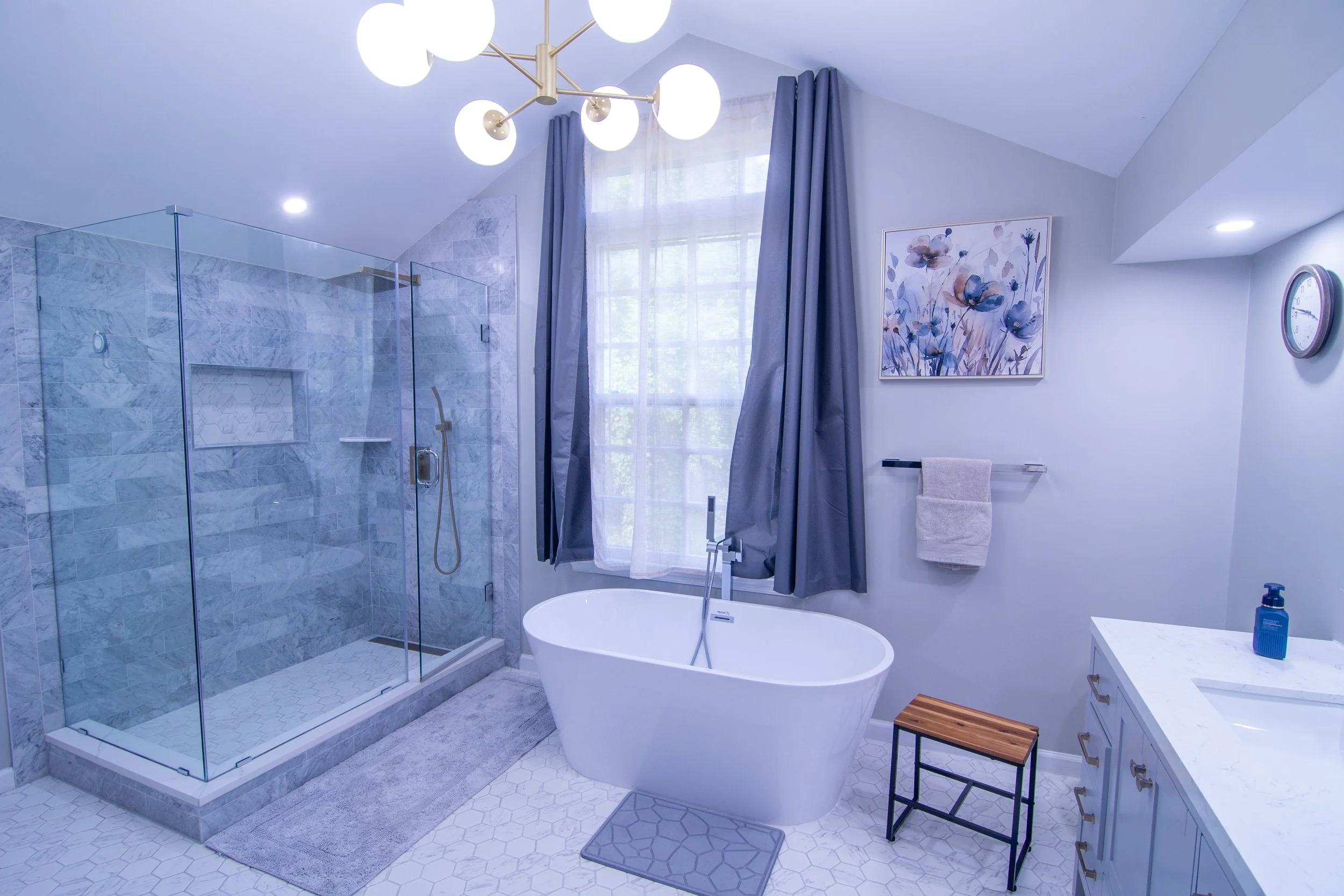 A modern bathroom with a glass-enclosed shower, a freestanding bathtub, a vanity with a sink, and a large window with blue curtains. Decor includes a floral painting, a wall clock, a towel rack with towels, and a chandelier with globe lights.
