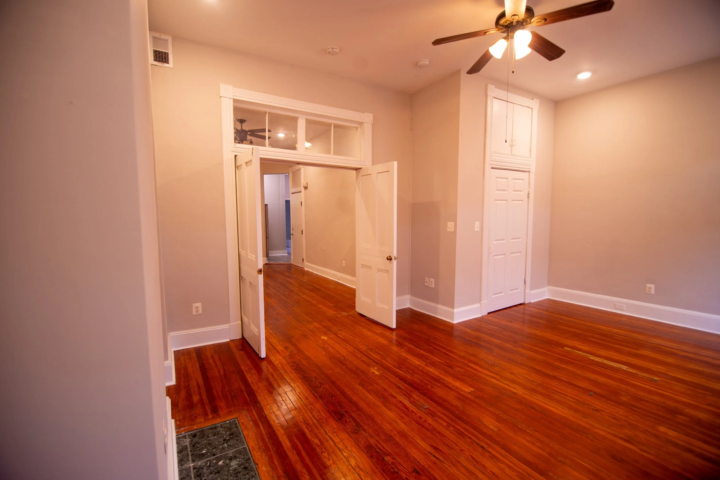 An empty living room with hardwood floors, beige walls, and white trim. There is a ceiling fan with lights, a door leading to another room, and what appears to be a closet with white doors.