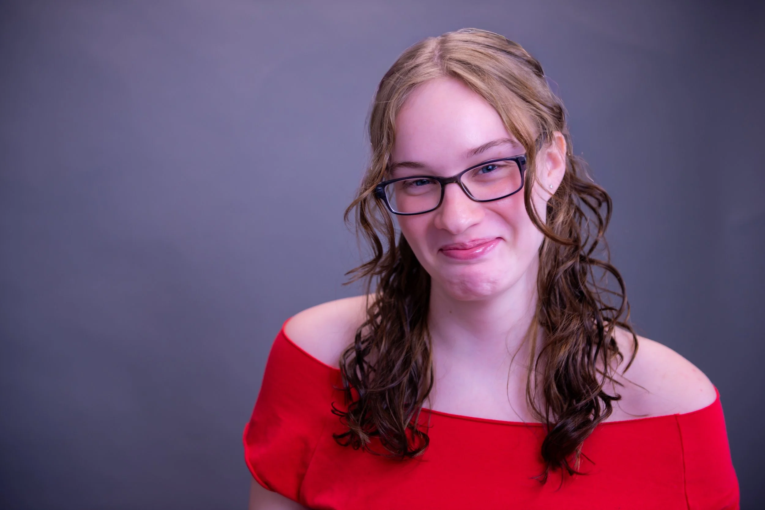 Young woman with wavy red hair wearing glasses and a red off-shoulder top, smiling slightly, against a plain gray background.