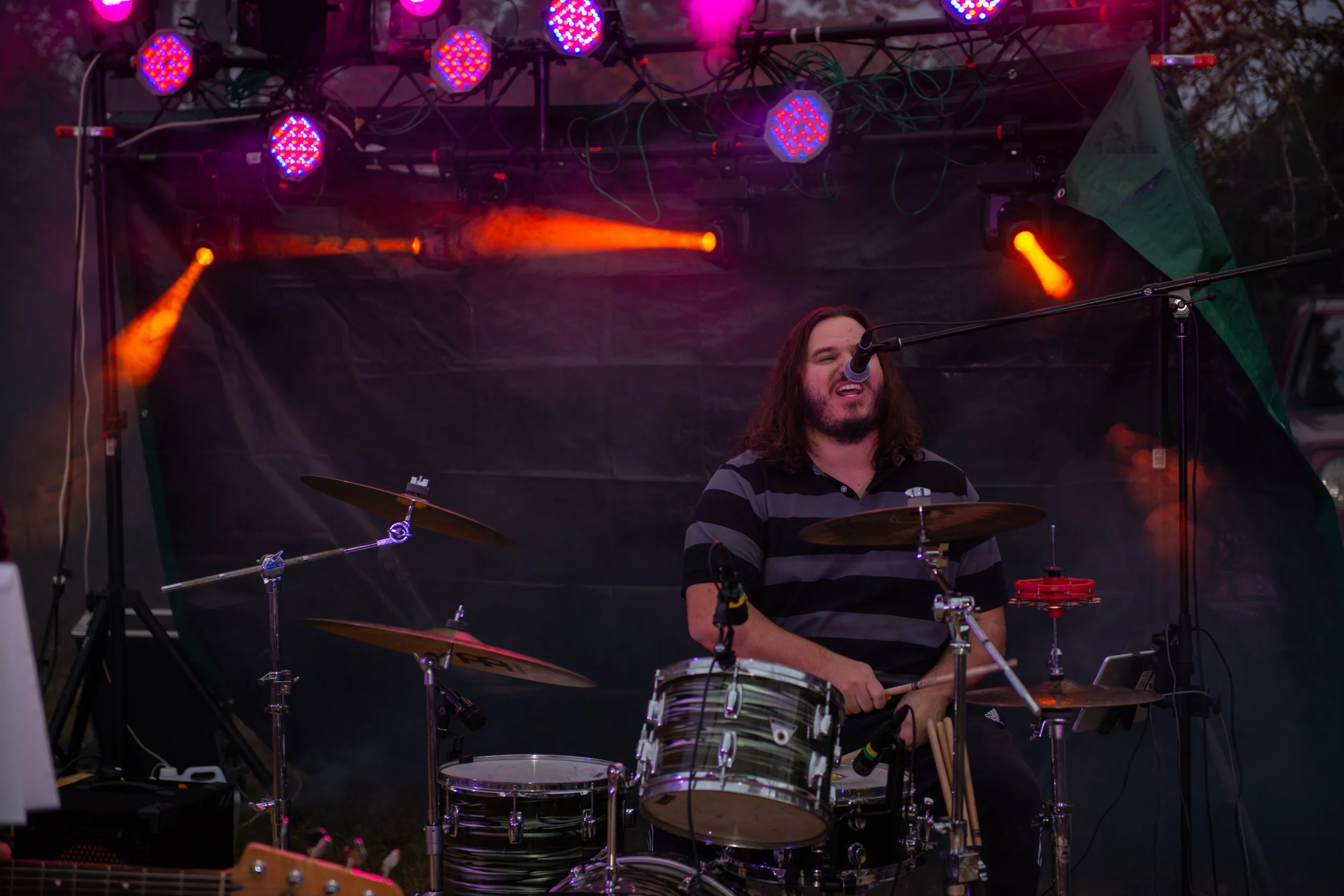 A male drummer with long hair and a striped shirt performing on an outdoor stage with colorful stage lights.