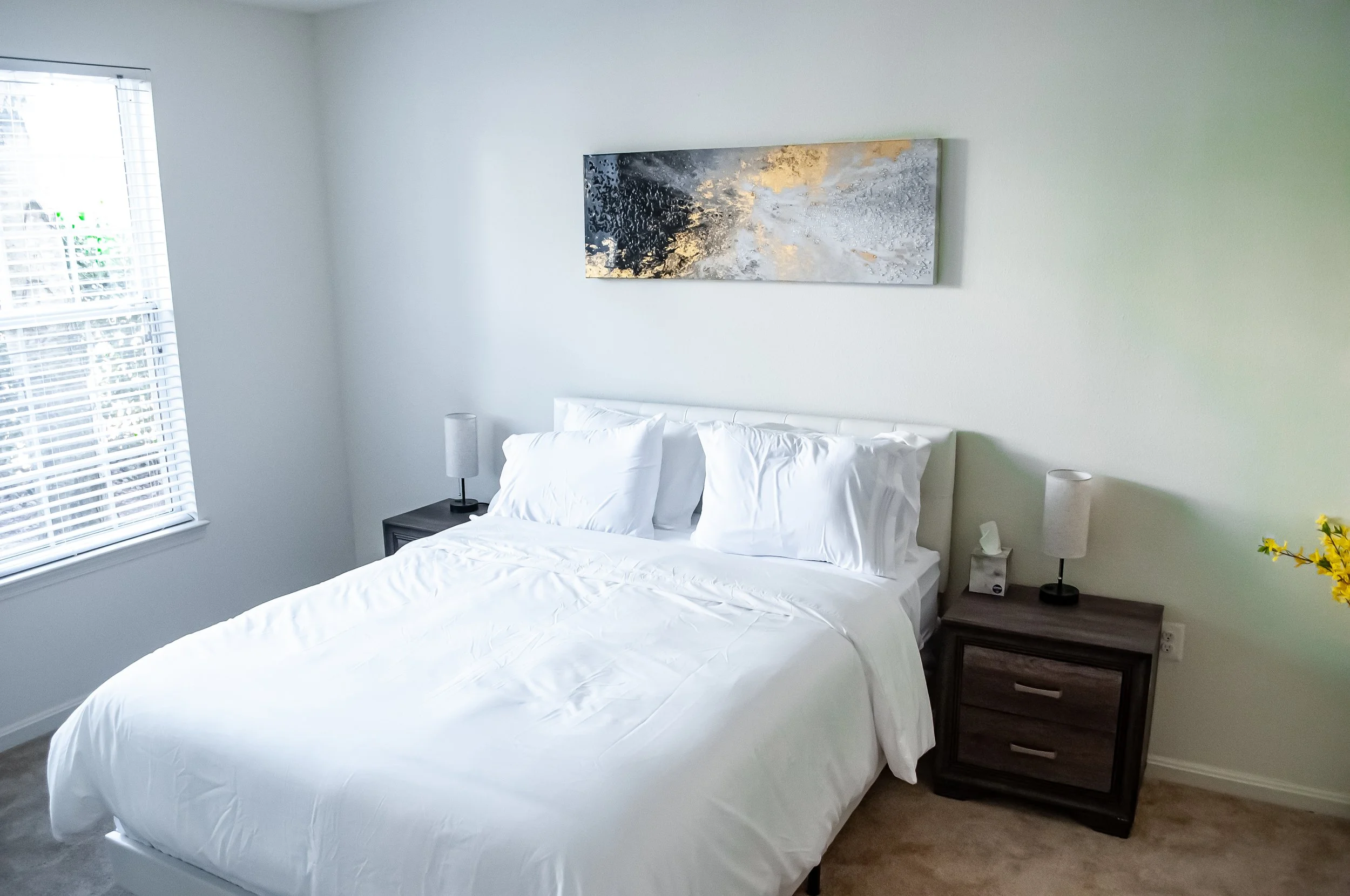 A bedroom with a bed, white bedding, and a nightstand with a lamp on each side, a window with blinds, a piece of abstract wall art, and a small yellow flower in the corner.
