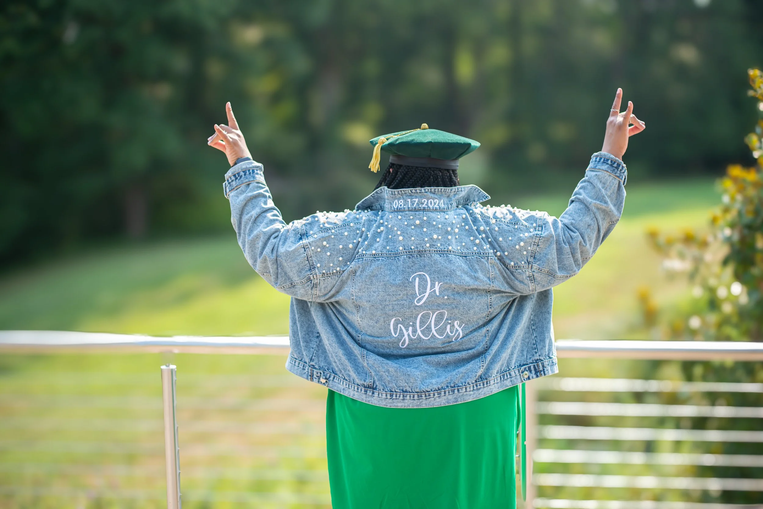 Back view of a person wearing a denim jacket with 'Dr. Gillis' embroidered on the back, a graduation cap, and green pants, standing outdoors with arms raised, pointing upwards, in a park with blurred green trees in the background.