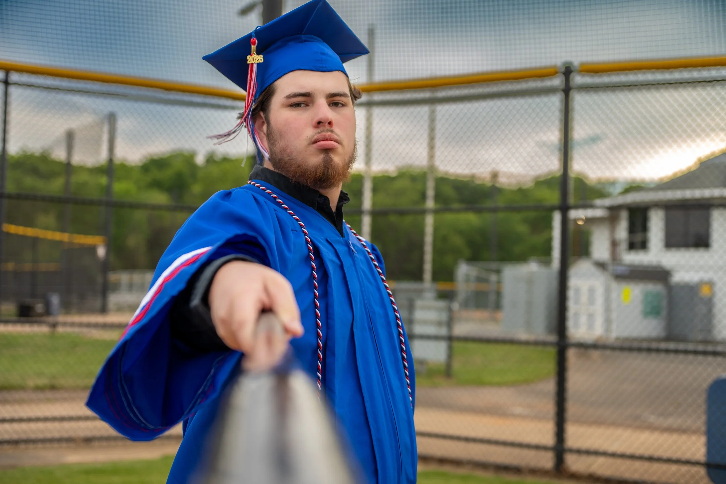 A young man in a blue graduation gown and cap with a '2026' tassel stands on a baseball field, holding a baseball bat.